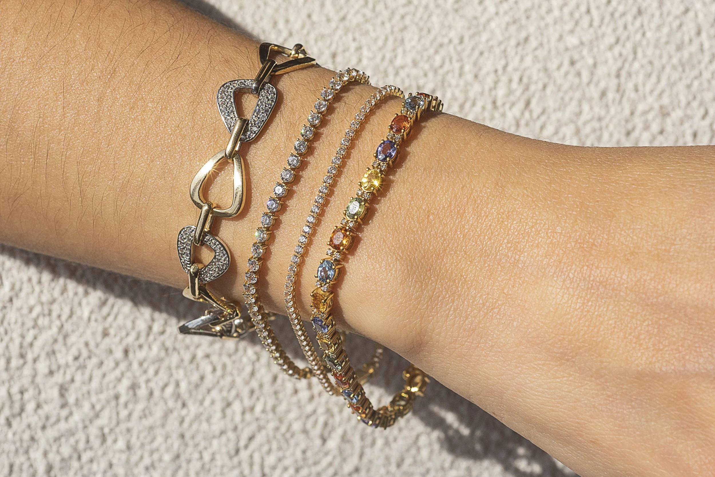 Muñeca con cinco pulseras de diferentes diseños y colores, con superficie de arena o piedra clara en el fondo.