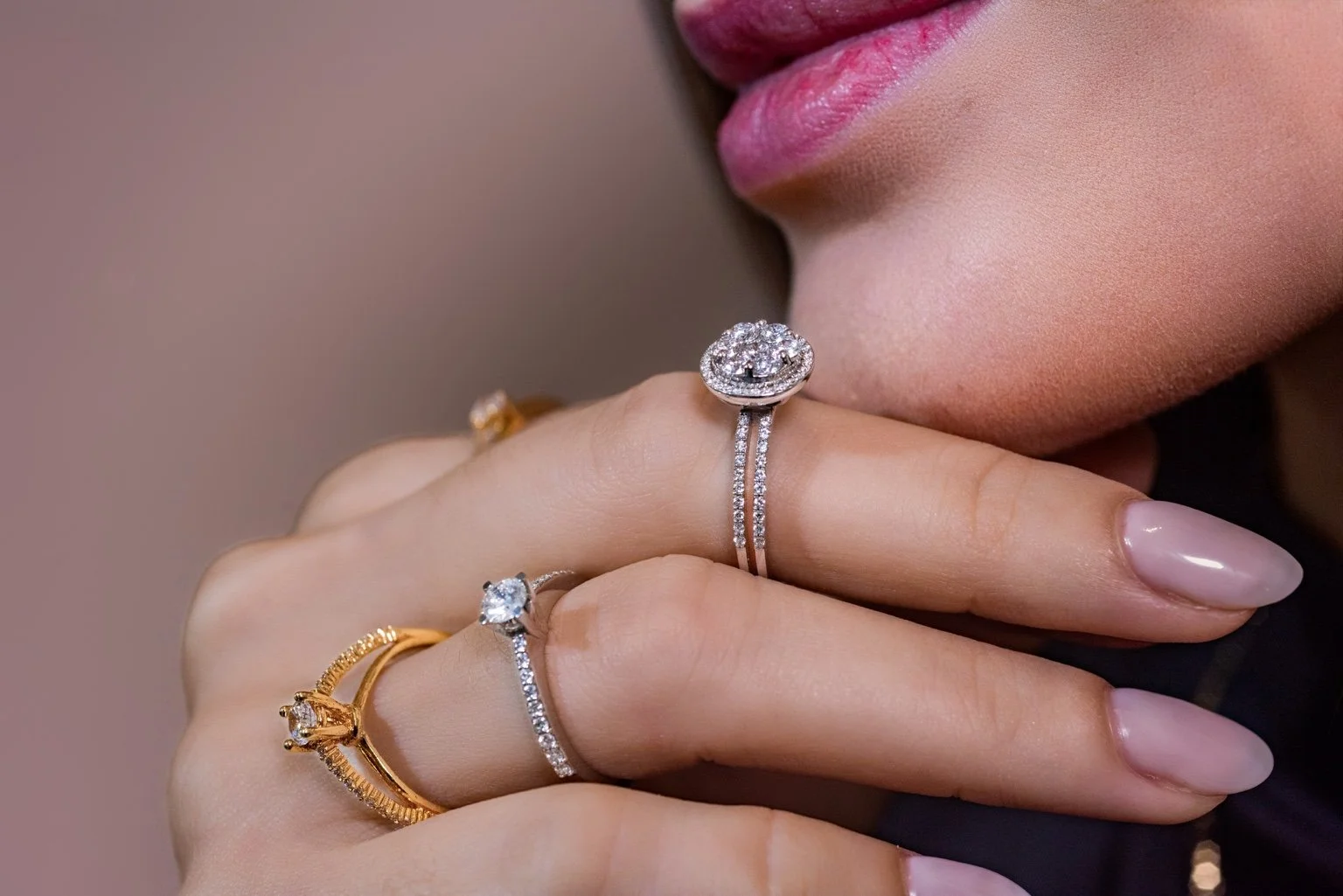 Manos con varios anillos con diamantes y oro, cerca del rostro de una mujer con labios rosados, en un fondo difuso.