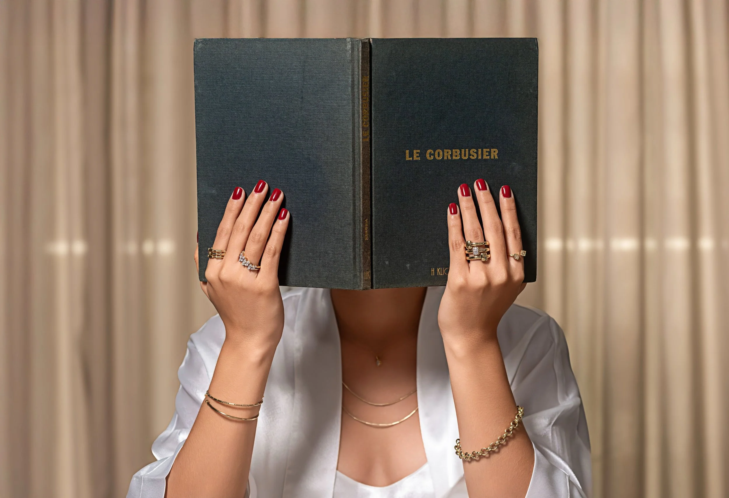 Persona con uñas rojas sostiene un libro titulado 'Le Corbusier' cubriéndola de la vista, usa múltiples anillos y pulseras, con fondo de cortinas beige.