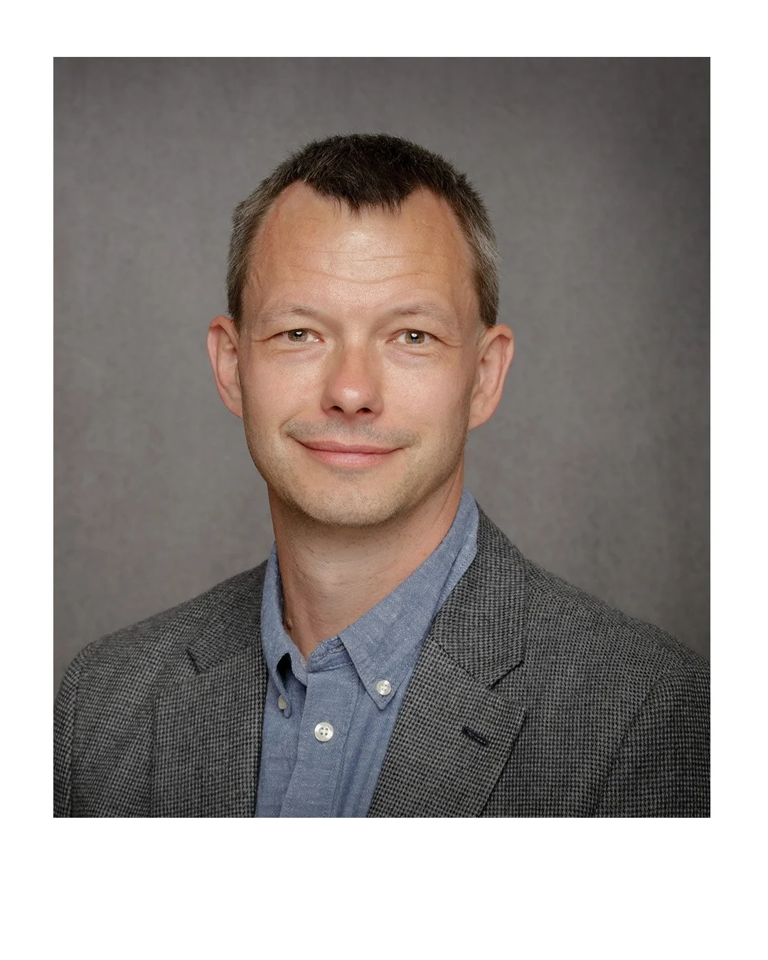 Headshot of a man with short brown hair, wearing a gray plaid blazer over a blue button-up shirt, smiling against a gray background.