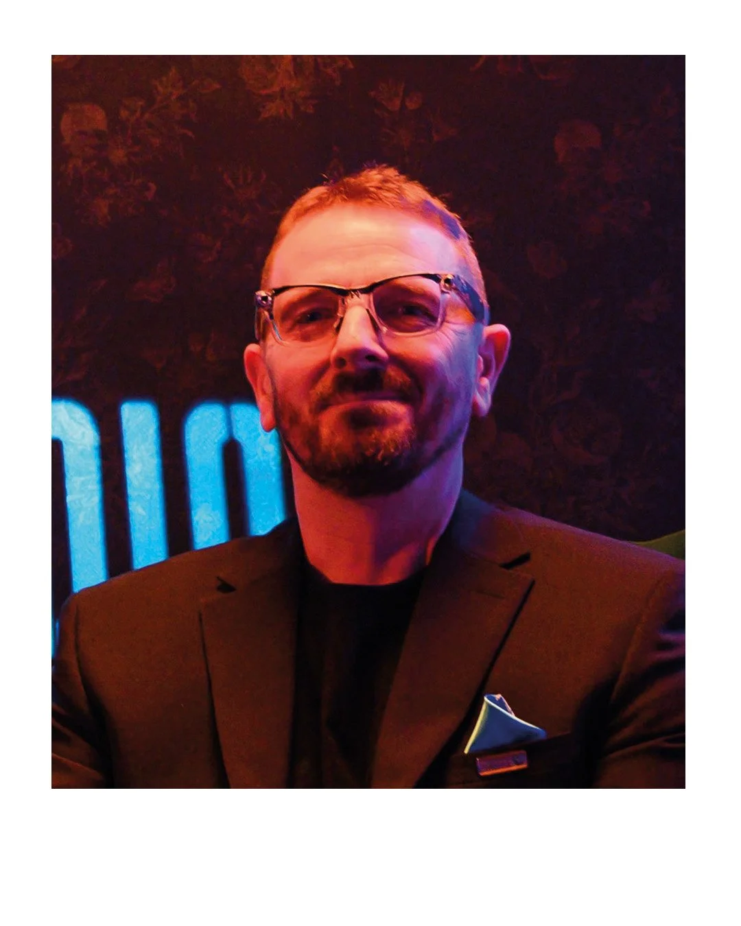 A man with a beard and glasses wearing a black blazer and black shirt, seated in a dimly lit environment with colorful lighting and a patterned background.