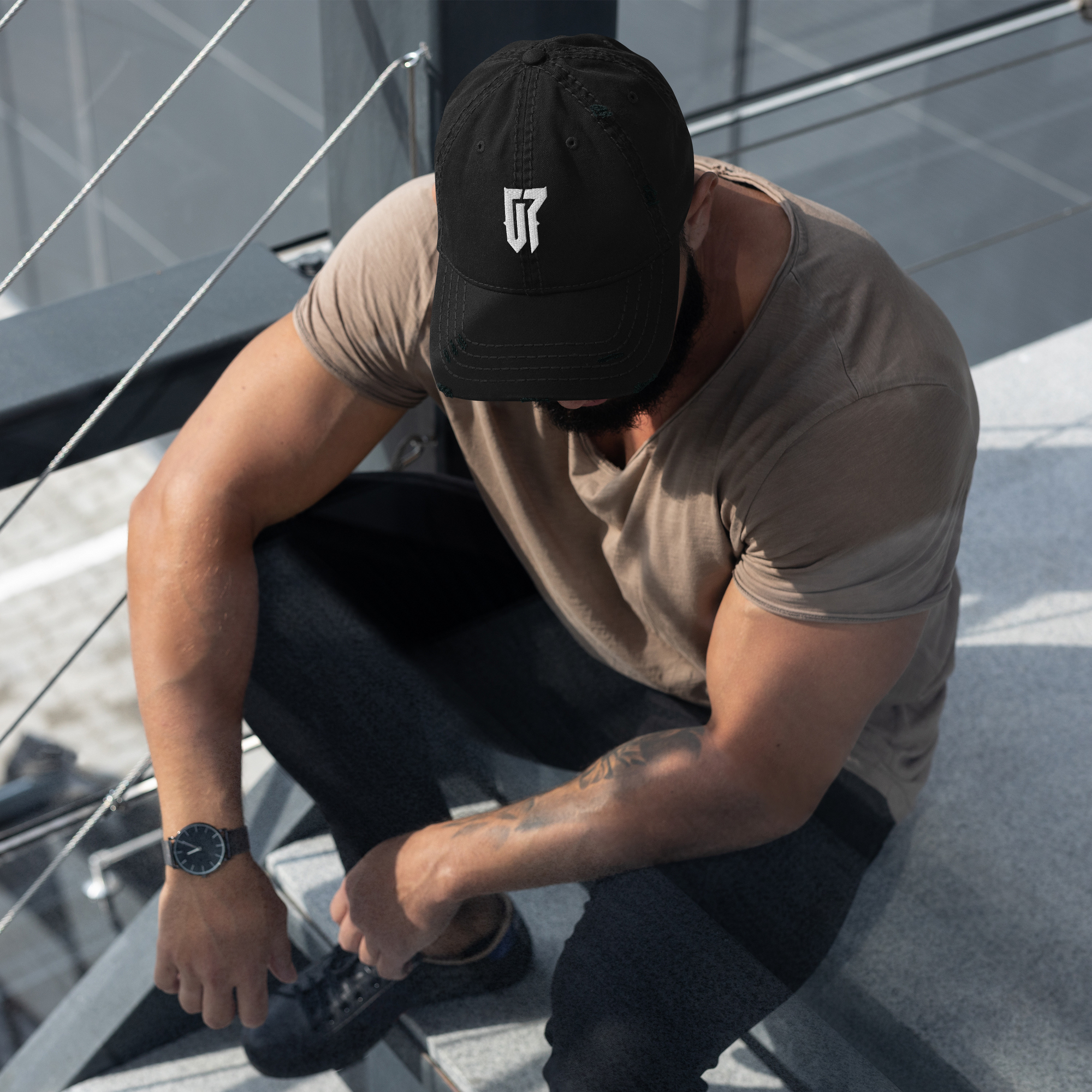 A man sitting on a ledge, looking down and tying his shoelace. He is wearing a black cap with a G7 Logo on it and a beige T-shirt, with a watch on his left wrist and tattoo on his right arm. www.onersn.com