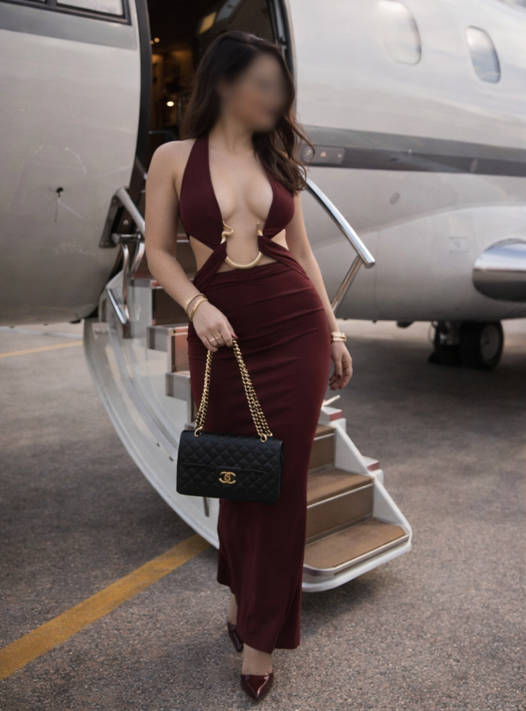 A woman in a burgundy dress with cutouts and gold accents is descending the stairs from a private jet, holding a black Chanel handbag with a gold chain strap.