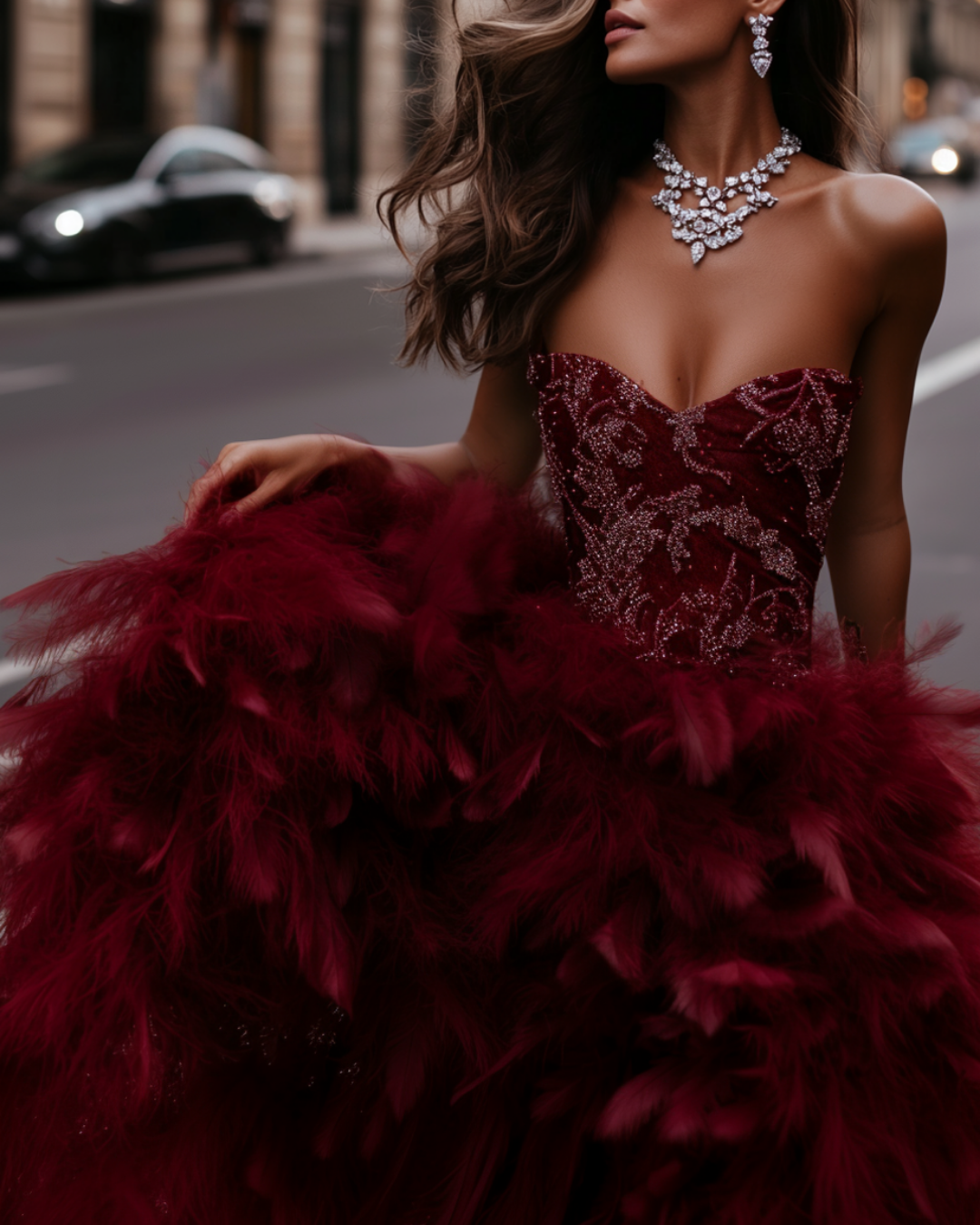 A woman in a strapless red gown with intricate beadwork, holding up a feathered skirt on a city street.
