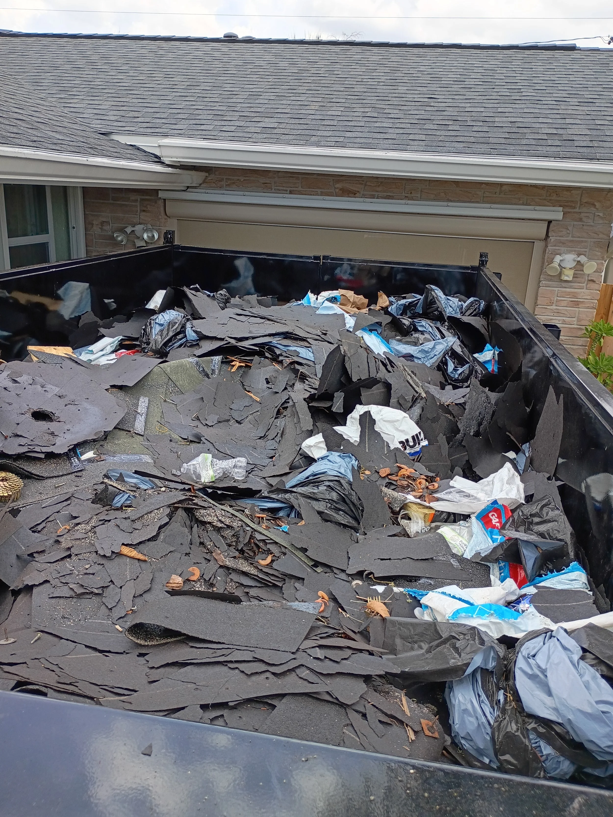 Top view of a construction or debris dump truck filled with torn black roofing shingles, plastic waste, and discarded items, with a residential house and garage in the background.
