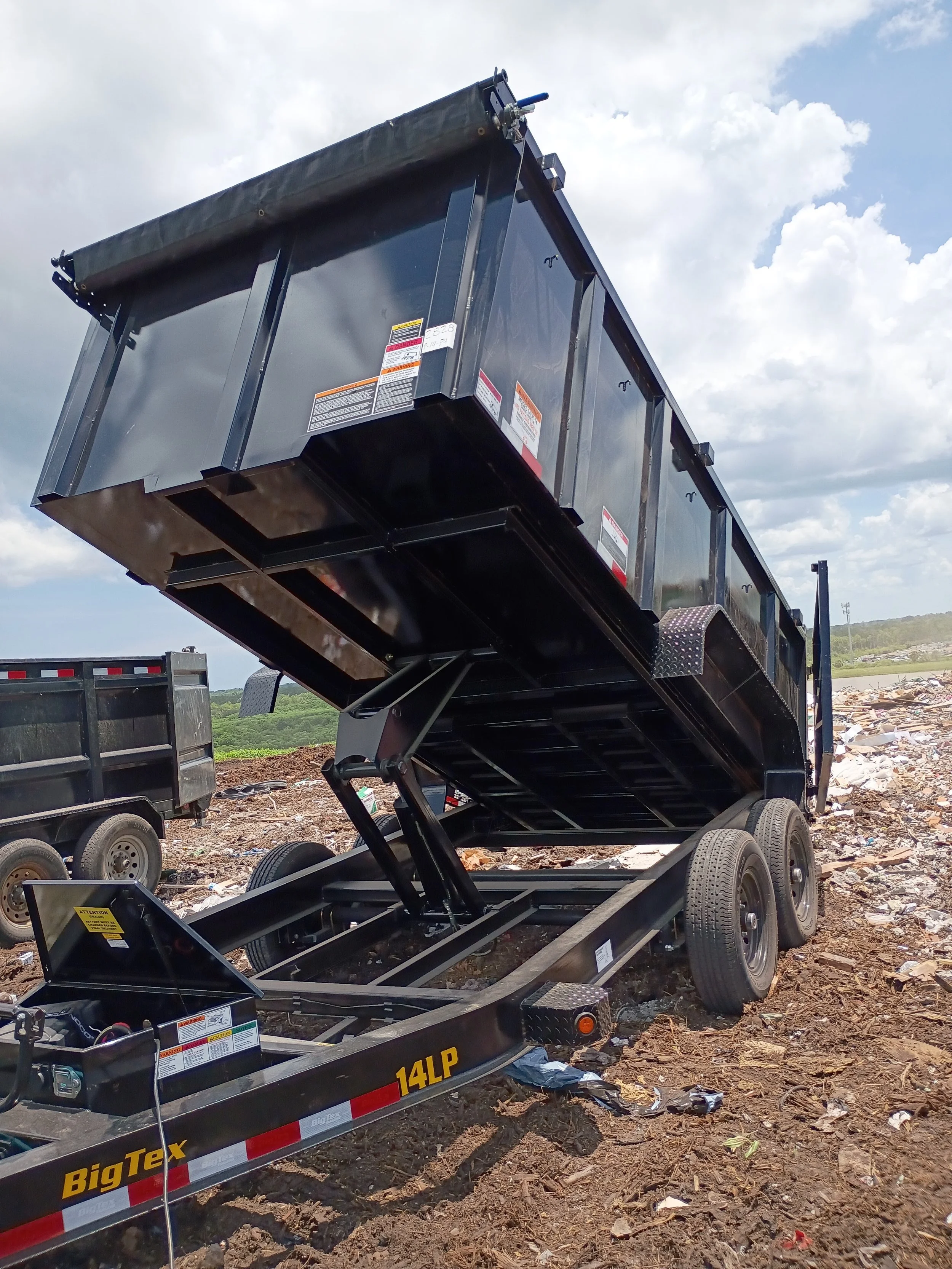 Black dump trailer with raised bed on a dirt lot, with a partly cloudy sky in the background.