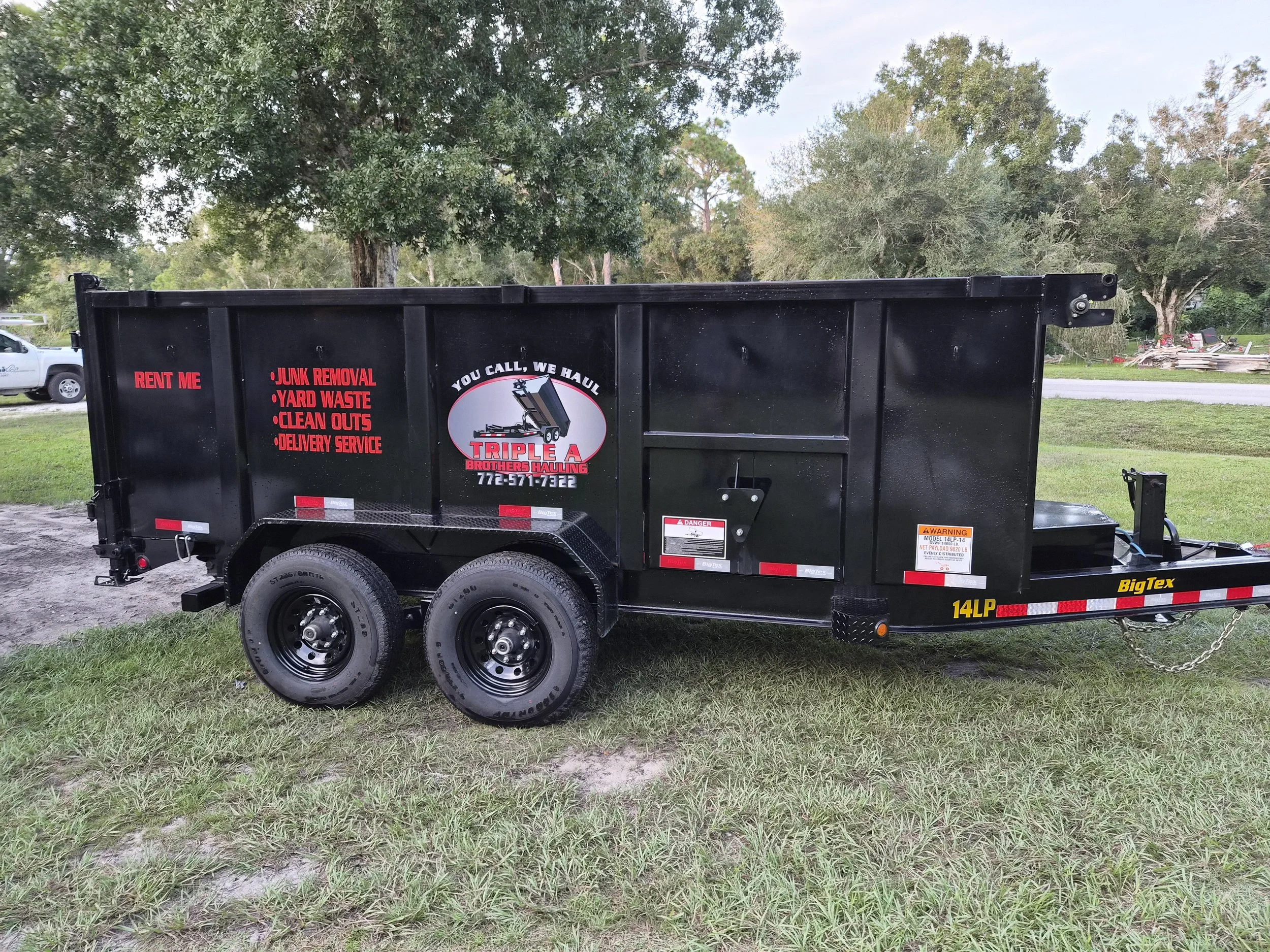 Black utility trailer with red and white text advertising Triple A Brothers Hauling, parked on grassy area with trees in background. The trailer has double wheels and a hitch, with various labels and warning signs.