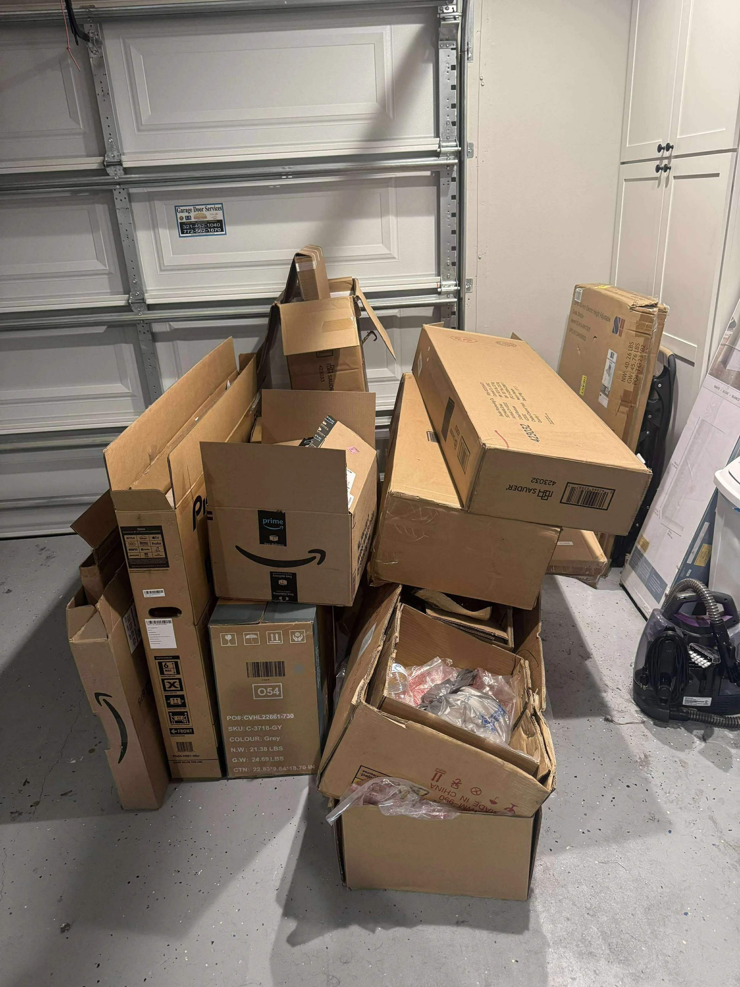 Several cardboard boxes stored in a garage, with a closed garage door in the background and a vacuum cleaner to the right.