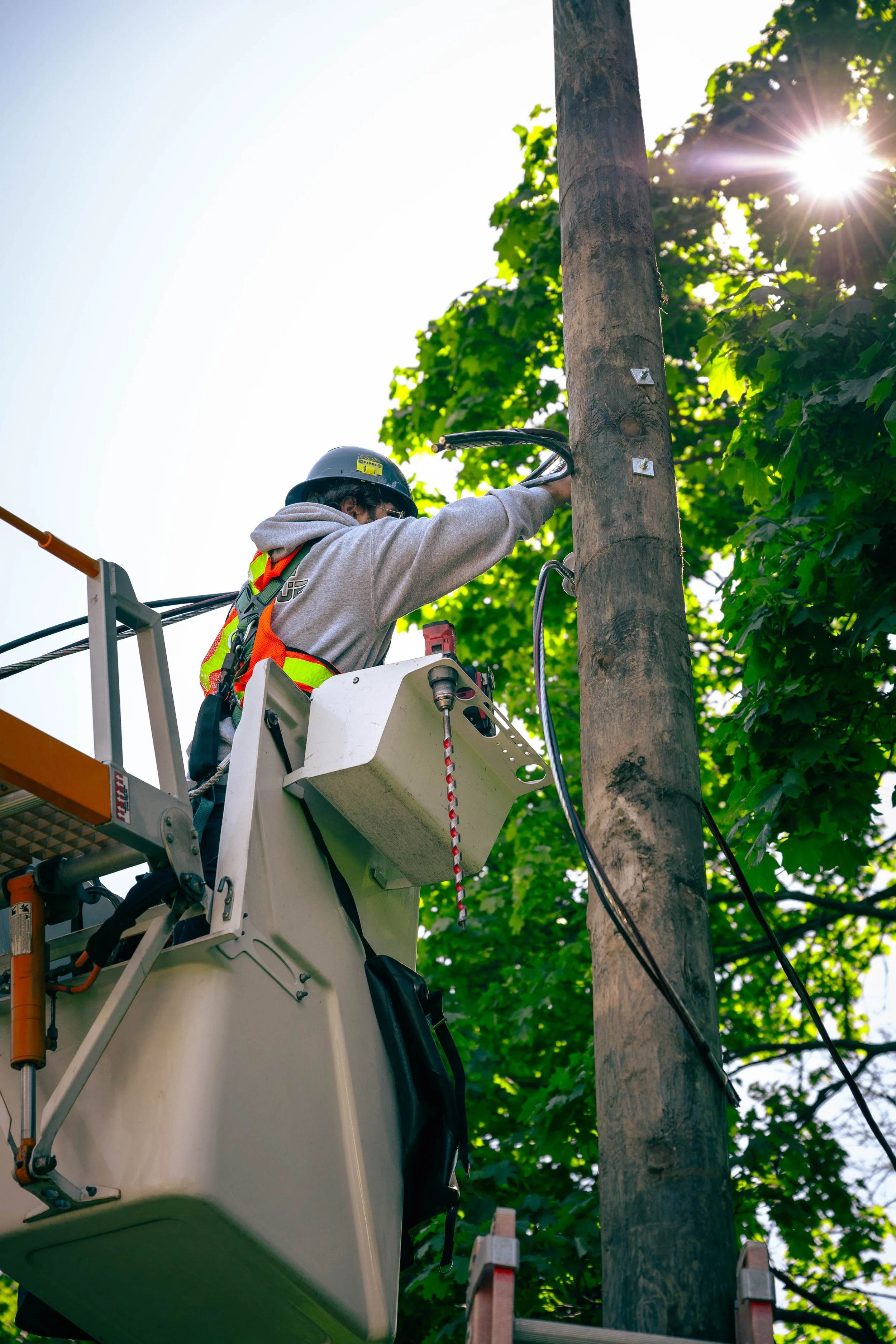 Electrician working on a utility pole from a cherry picker, with green trees and sunlight in the background.