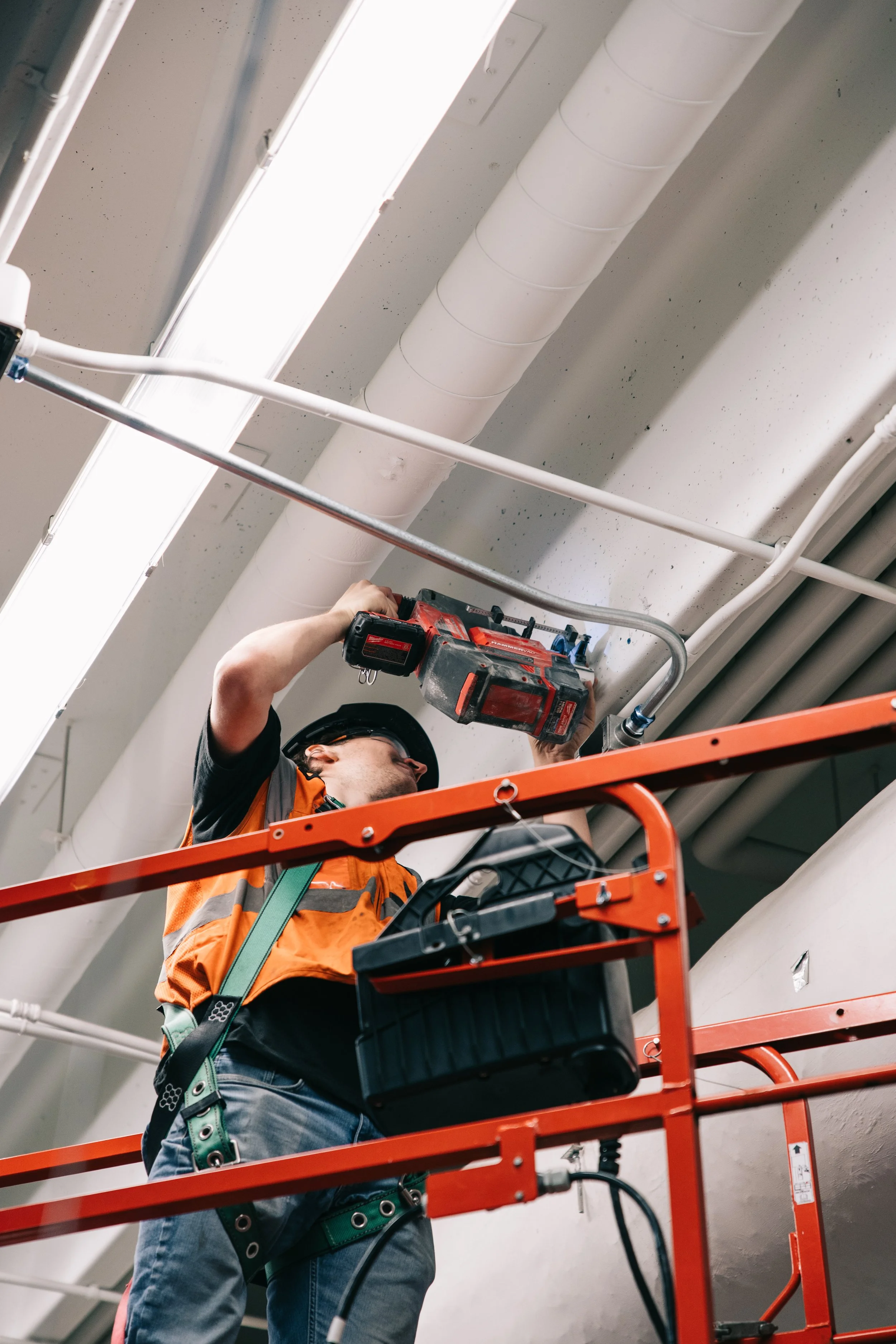 A construction worker in an orange safety vest and black hard hat is repairing or installing wiring on the ceiling of a building, using a power drill while standing on an orange scaffolding.
