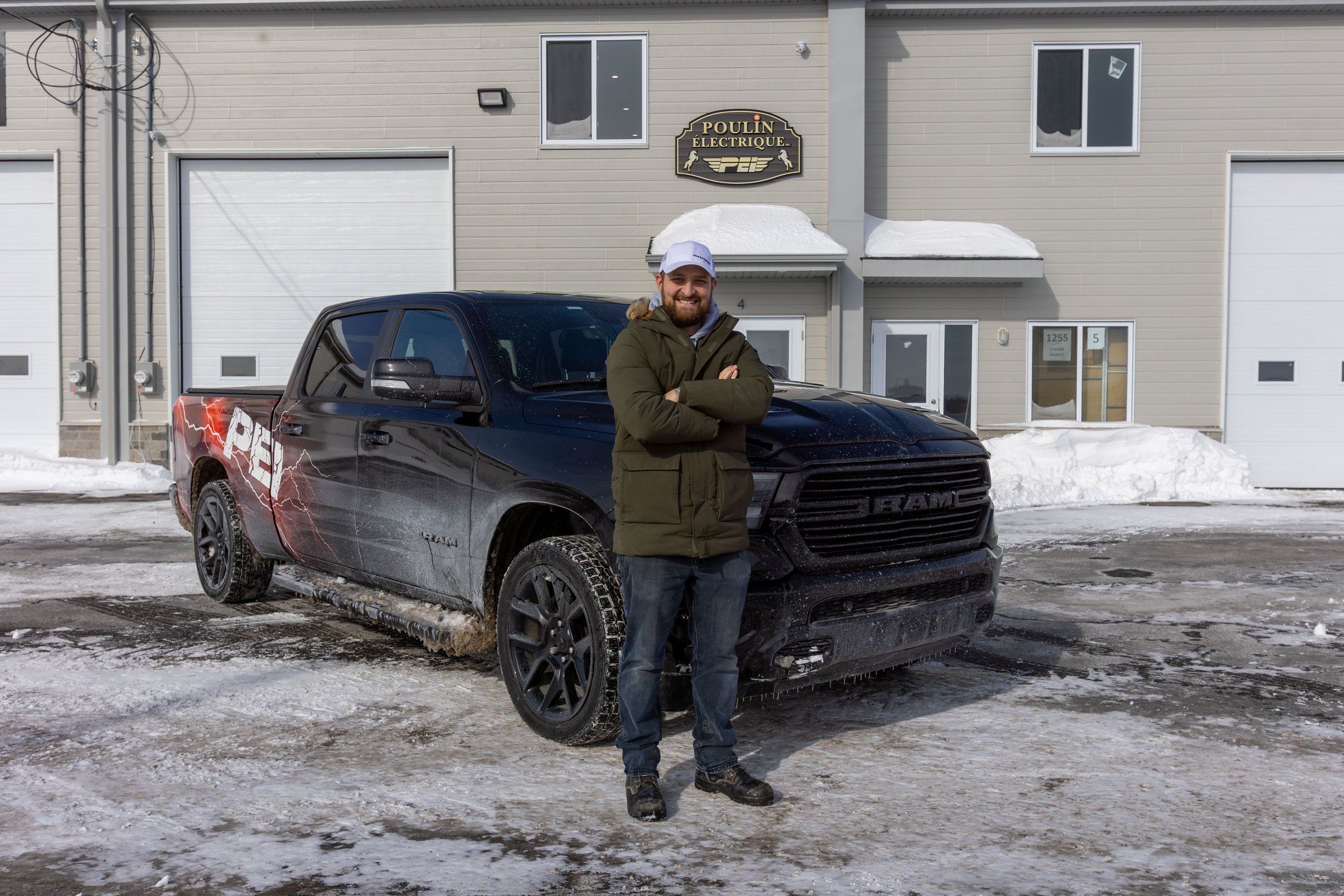 A man in winter clothing standing in front of a black RAM pickup truck outside a commercial building during winter. The building has a sign reading 'Poulin Électrique.' Snow is piled around the building and on the ground.