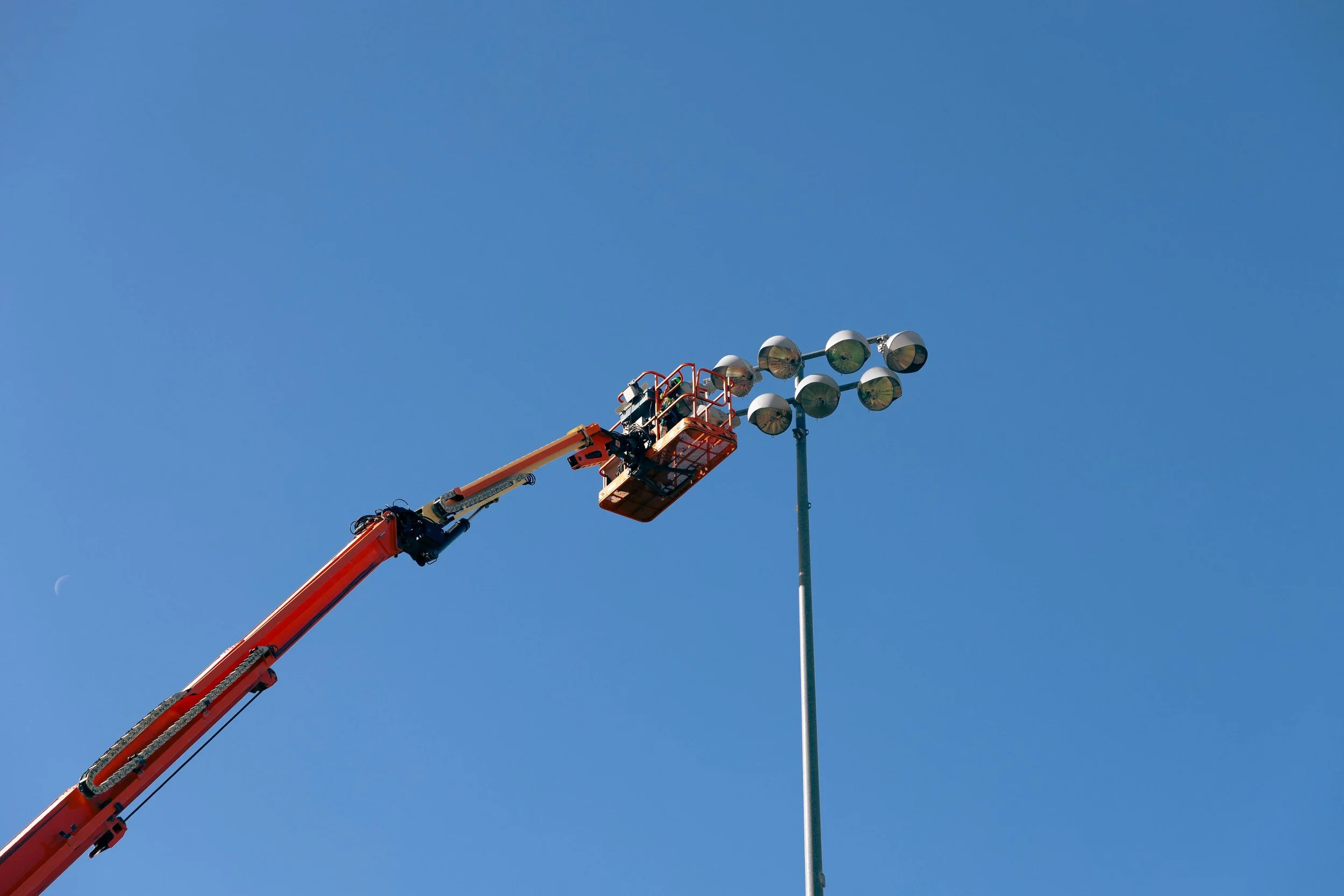 A crane lifting a worker to maintenance or inspection of stadium or flood lights on a tall pole against a clear blue sky.