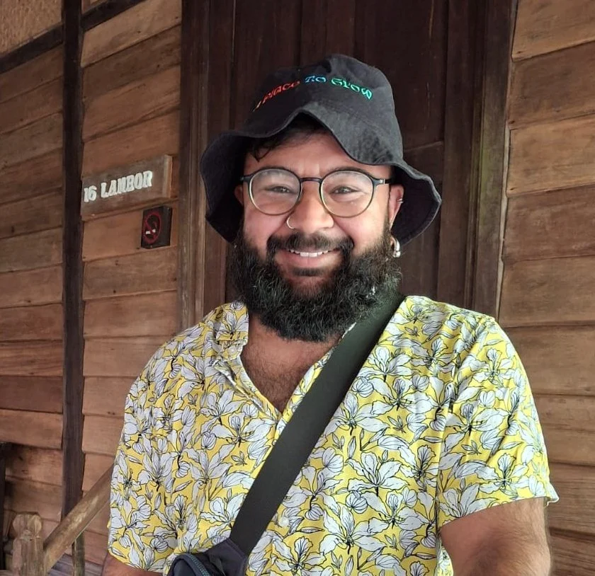Smiling man with a beard, glasses, hoop earrings, and a nose ring, wearing a black bucket hat with colorful text, a yellow floral shirt, and a black messenger bag strap, standing in front of a wooden wall with a sign reading '16 LABOR'.
