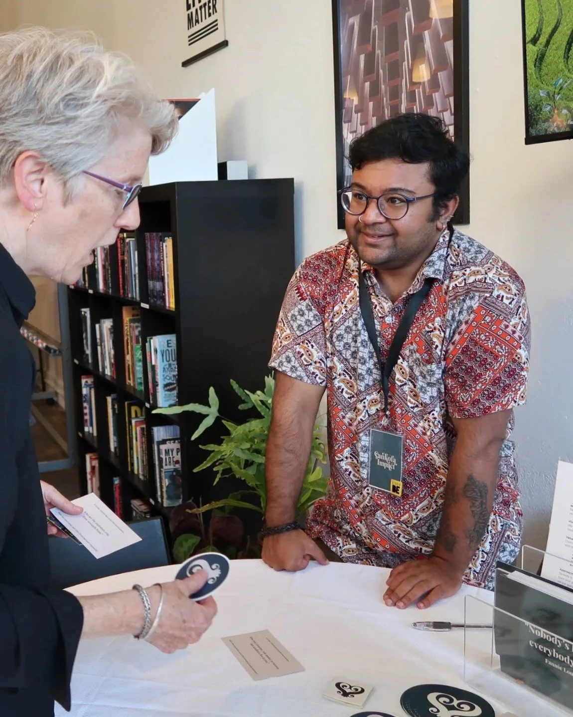 A young man with glasses and a patterned shirt is standing and leaning on a table, smiling and looking at a woman with gray hair and glasses who is holding a card and a token. They are in a bookstore or library with bookshelves and posters in the background.