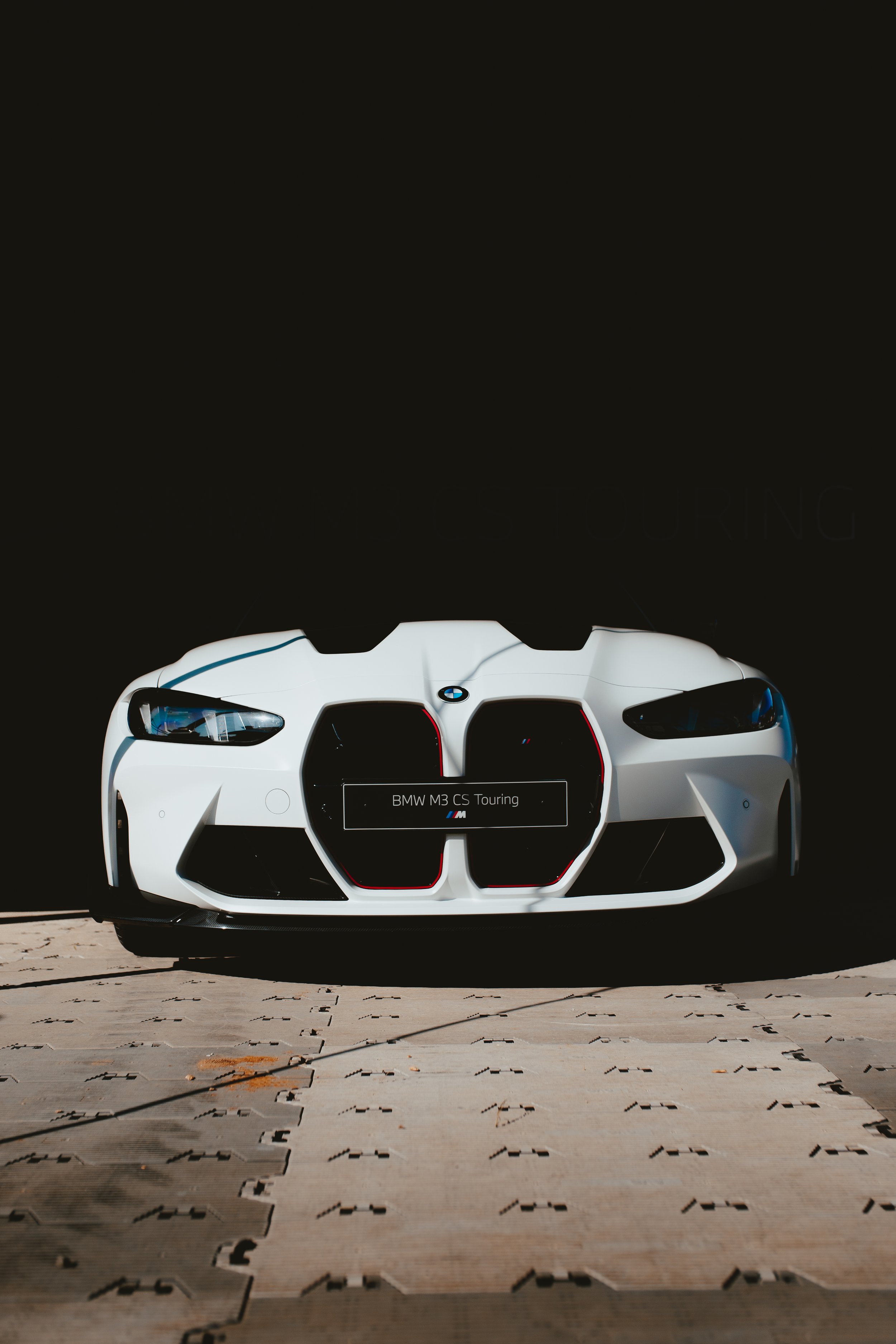 A white BMW M3 CS Touring car displayed on a platform with a black background.