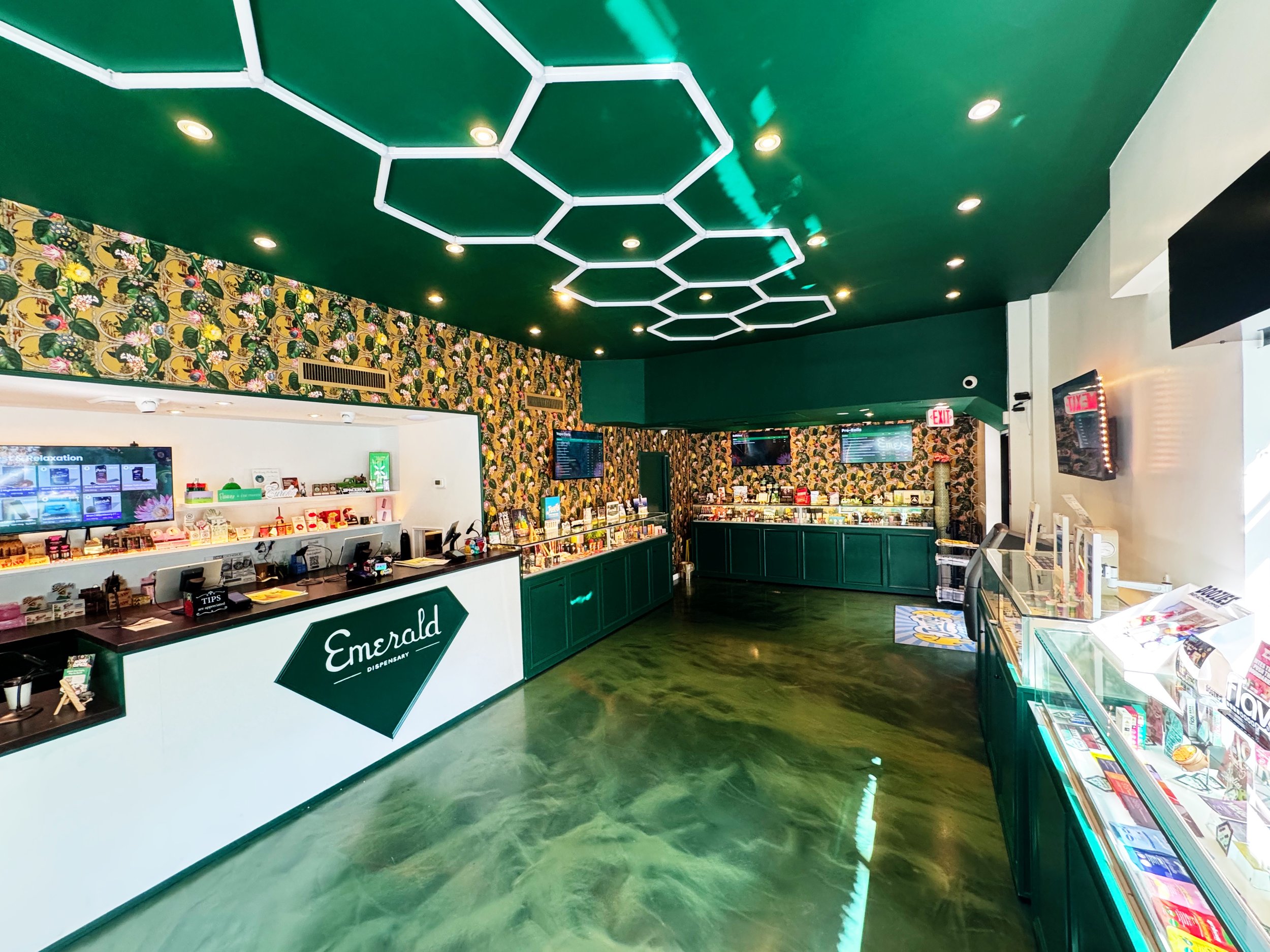 Interior of a dispensary with a white and green counter labeled Emerald Dispensary, decorated with a floral wallpaper, green ceiling with white hexagon lighting fixtures, and various glass display cases with product displays.