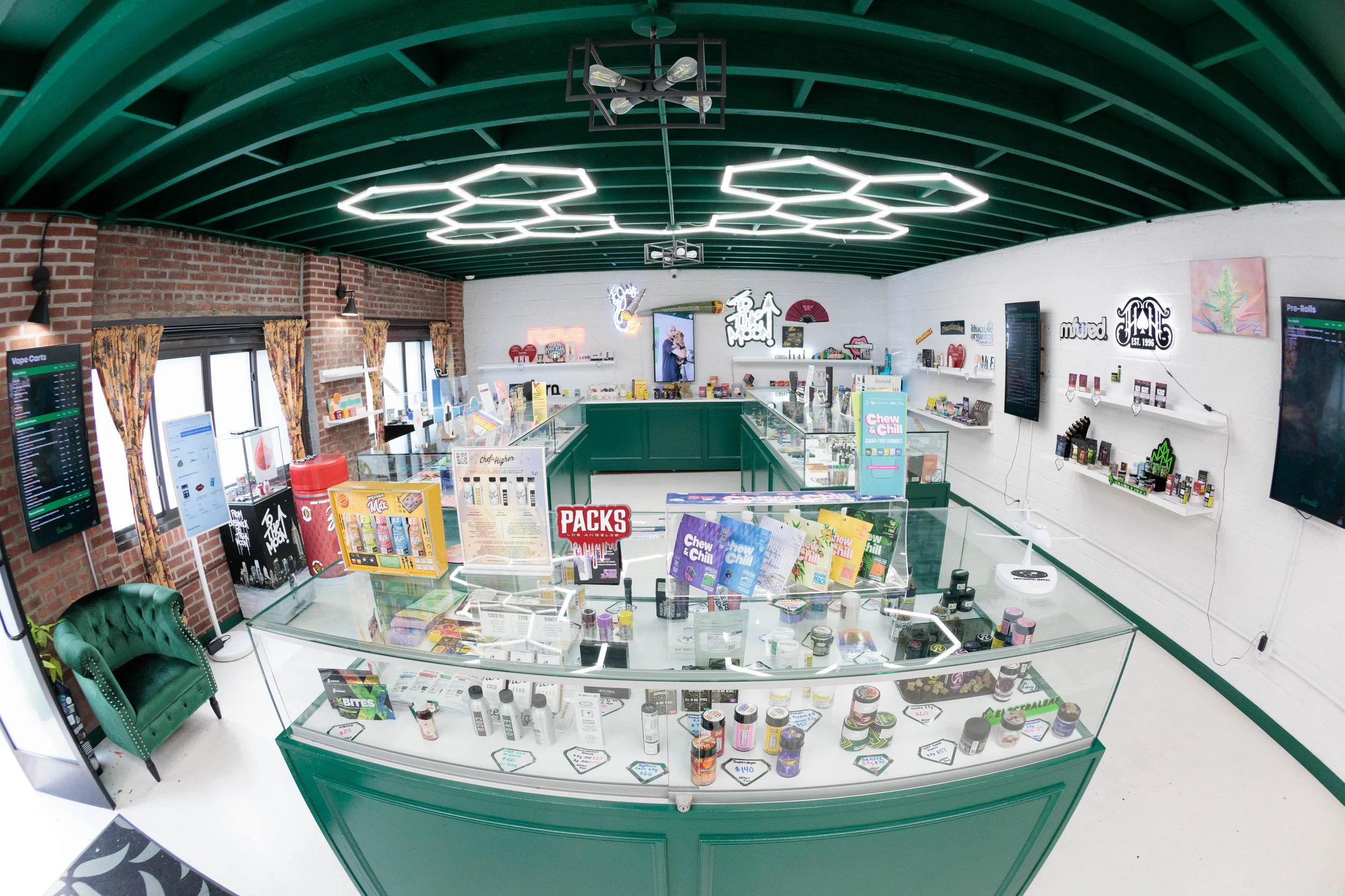 Interior of a modern vape shop with glass display cases filled with vaping products, shelves on the walls with more products, illuminated signs, a green velvet chair, and decorative lighting fixtures on the ceiling.