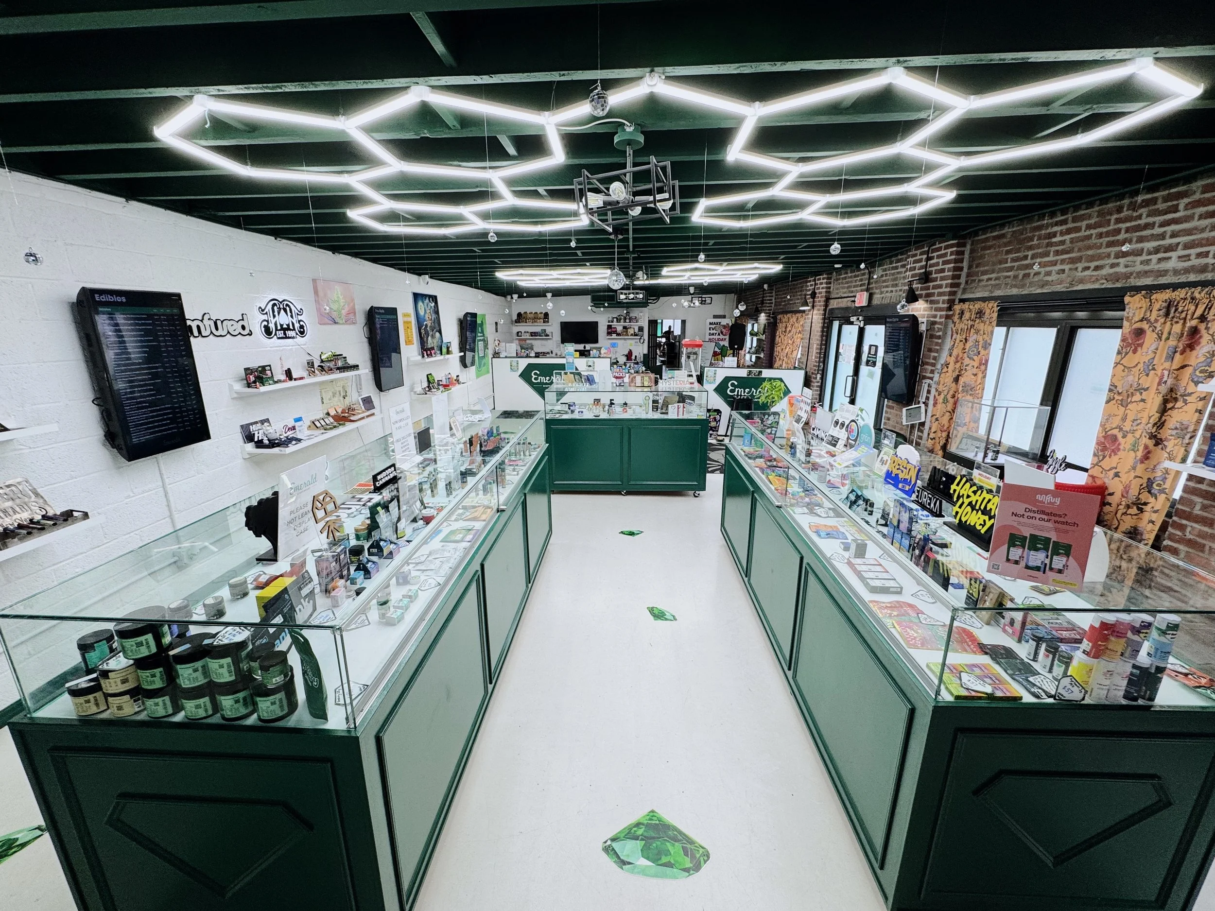 Interior of a retail store with glass display cases, electronic screens on white and brick walls, ceiling with geometric LED light fixtures, and shelves with various products.