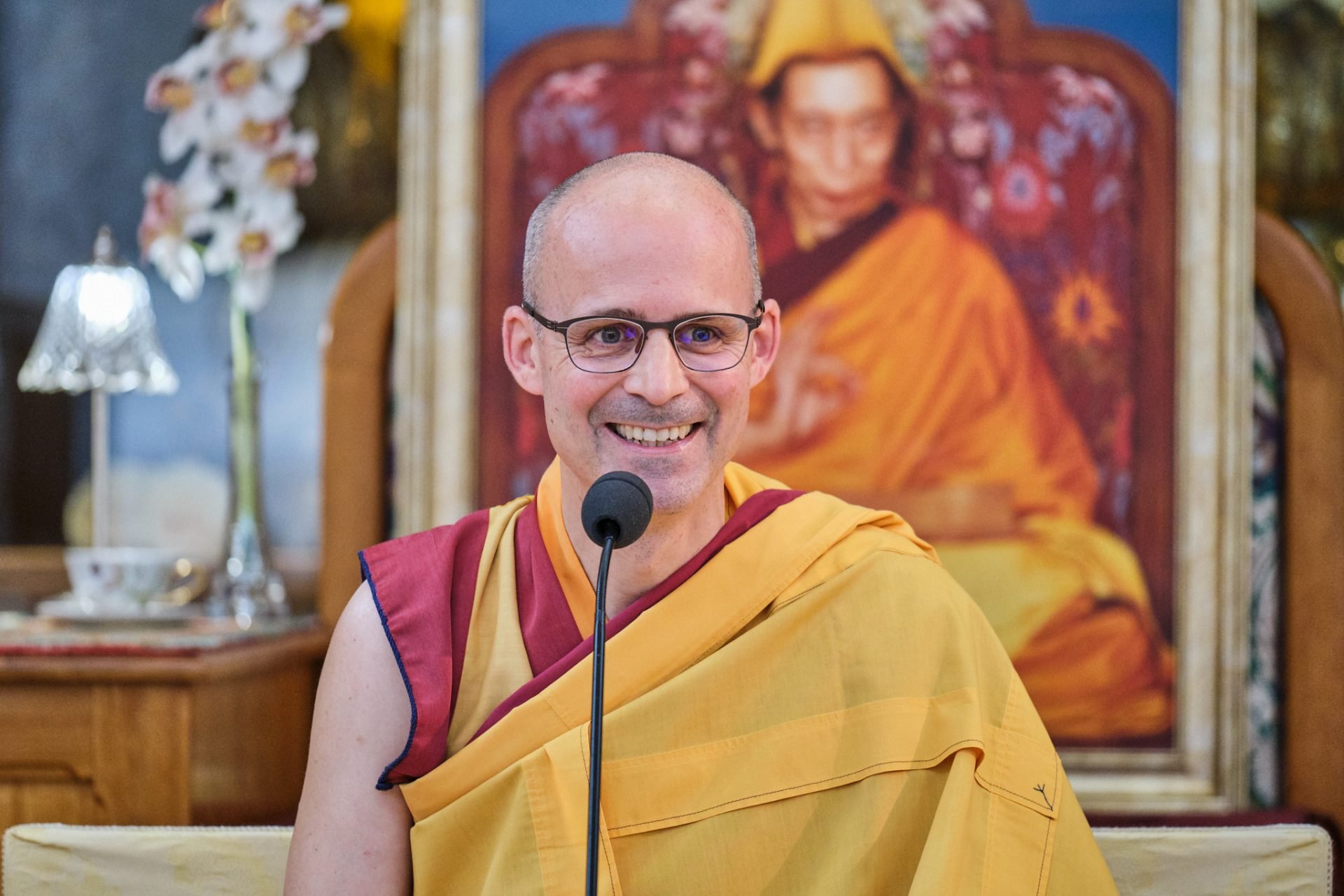 A smiling man wearing glasses dressed in traditional Buddhist monk robes, sitting in front of a large portrait of a monk dressed in orange robes and wearing a yellow hat.