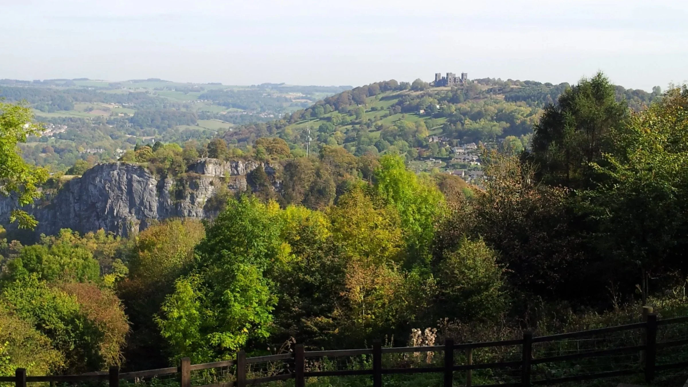 A panoramic view of green hills, a rocky cliff in the foreground, and a castle on a distant hilltop in the background.