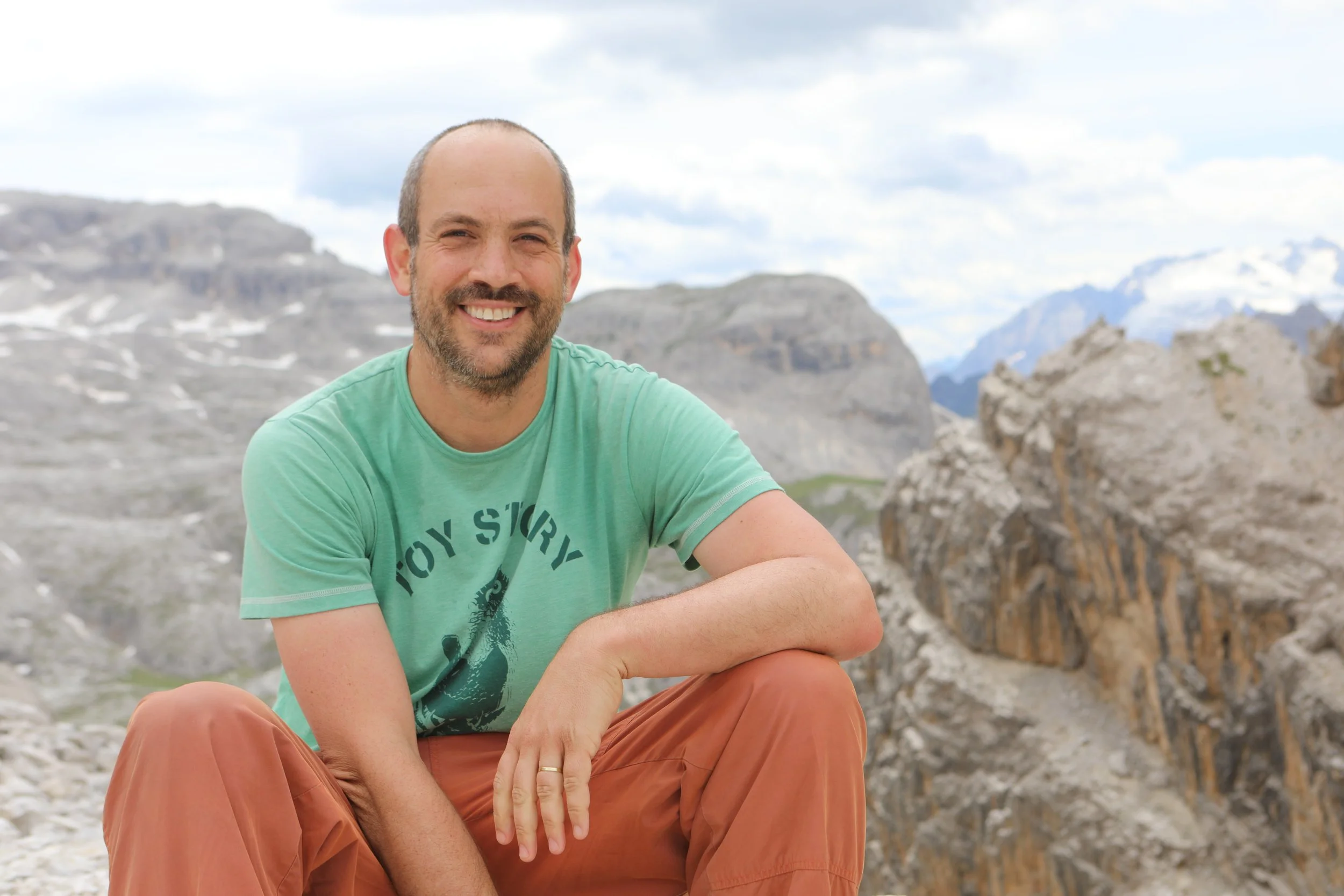 A man smiling outdoors with mountains in the background, wearing a green t-shirt and orange pants.