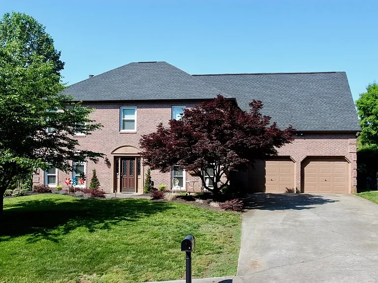 Front view of a two-story brick single family house with a gray roof, a driveway, and trees in the yard.