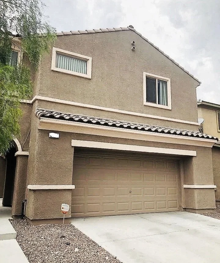 Two-story house with beige stucco exterior, tiled gable roof, two small windows on the upper floor, and an attached garage on the ground floor with a chain link fence at the driveway's edge.