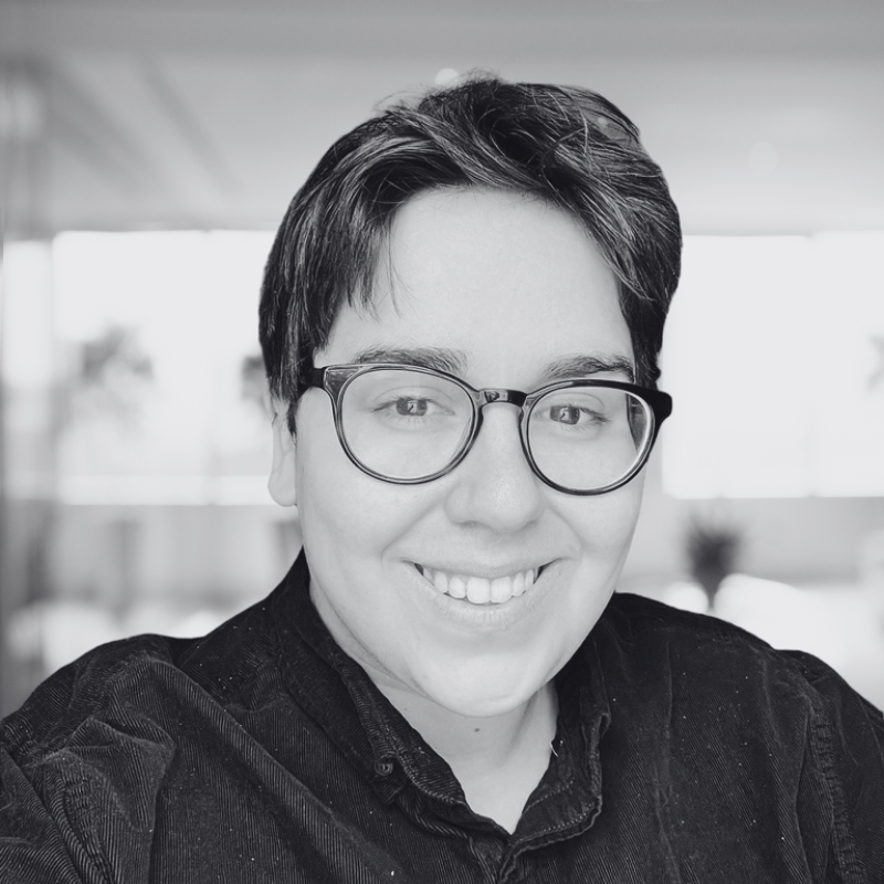 A smiling person with short dark hair, glasses, and a dark shirt indoors with window light and blurred background.
