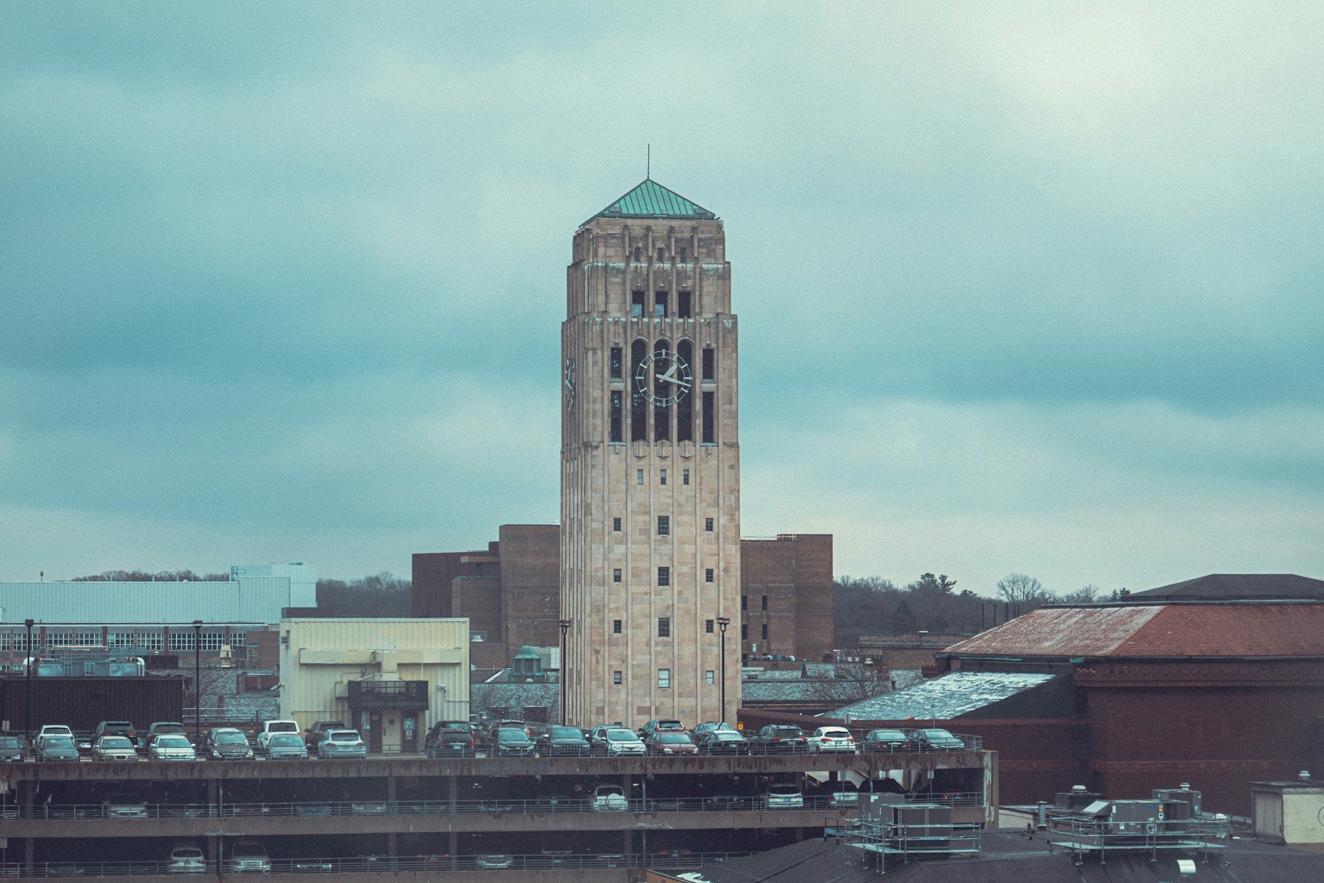 A cityscape featuring a tall clock tower with a green peaked roof and a large clock face, amid surrounding buildings and a parking lot with cars.