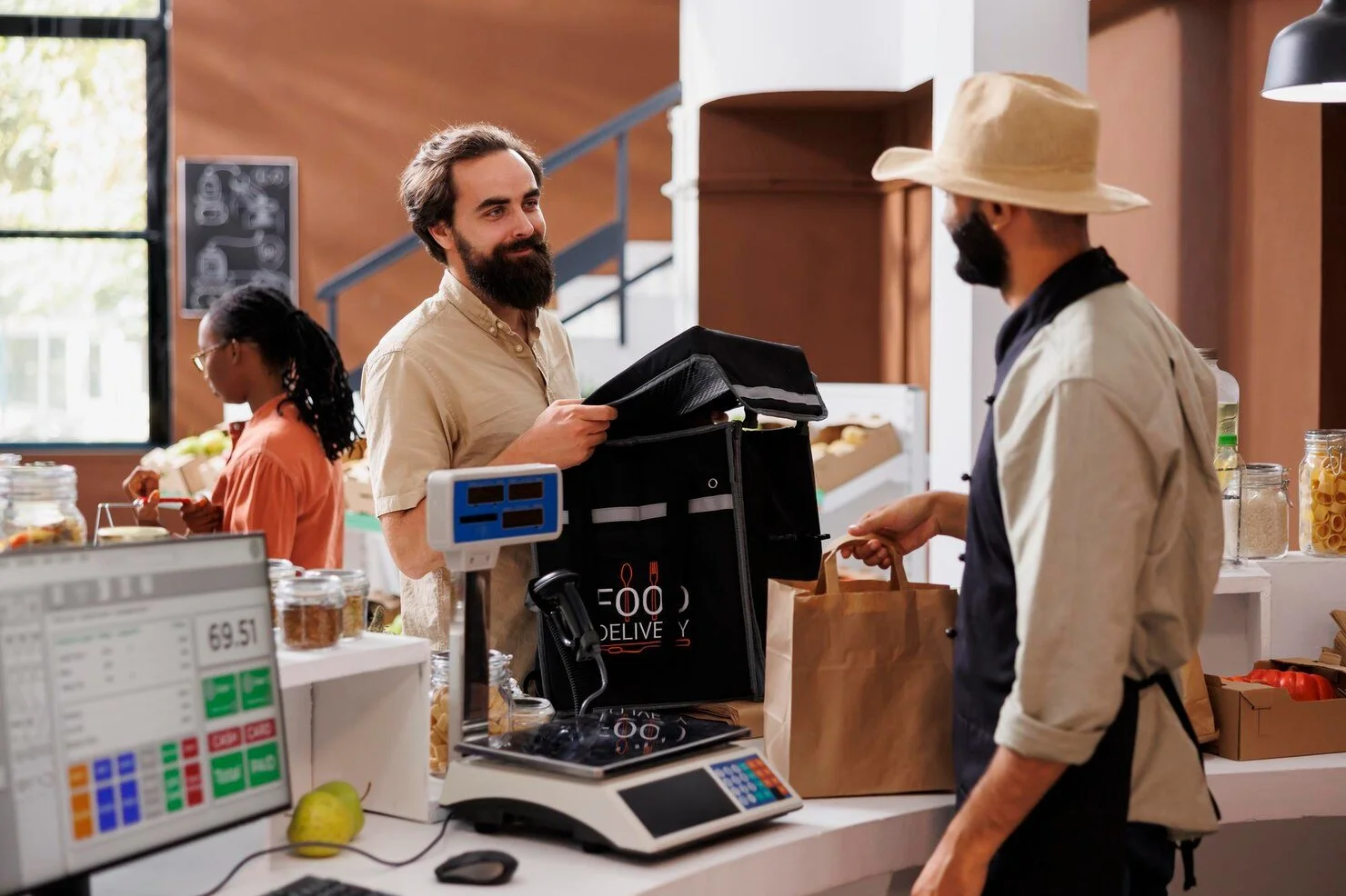 Man with beard and light-colored shirt shopping at a grocery store counter, talking to a worker in a hat and apron, while a woman in background shops.