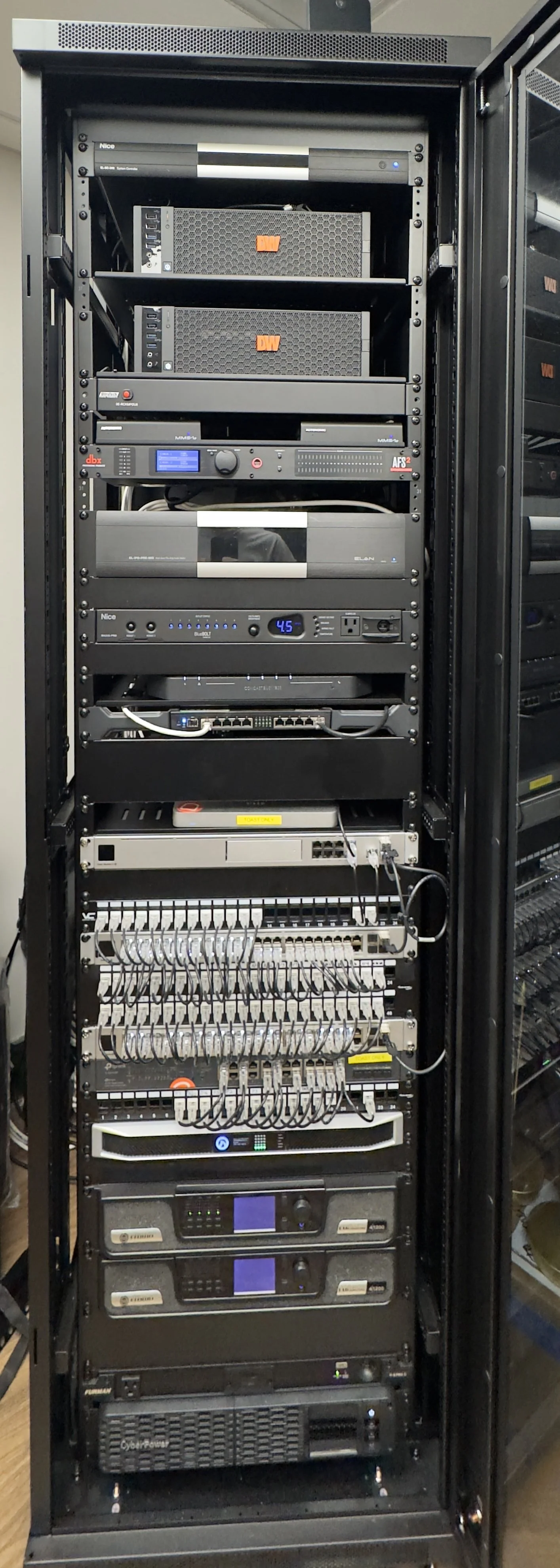 A tall black server rack containing multiple network and audio-visual equipment including switches, routers, amplifiers, and patch panels, with connected cables and LED indicator lights.