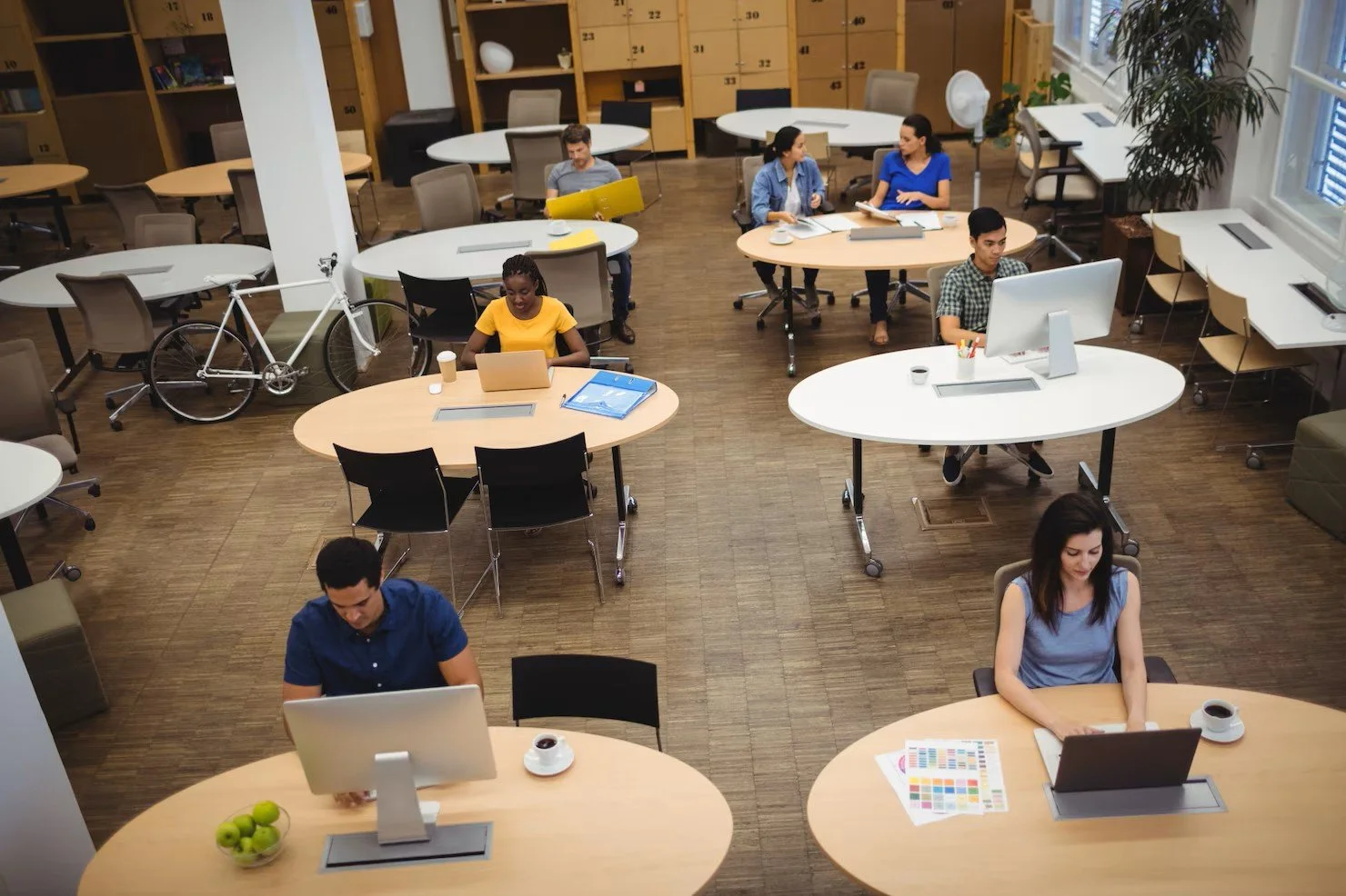 An office with multiple round tables and people working on computers. Some desks are empty, and there are potted plants, a bicycle, and windows with natural light.