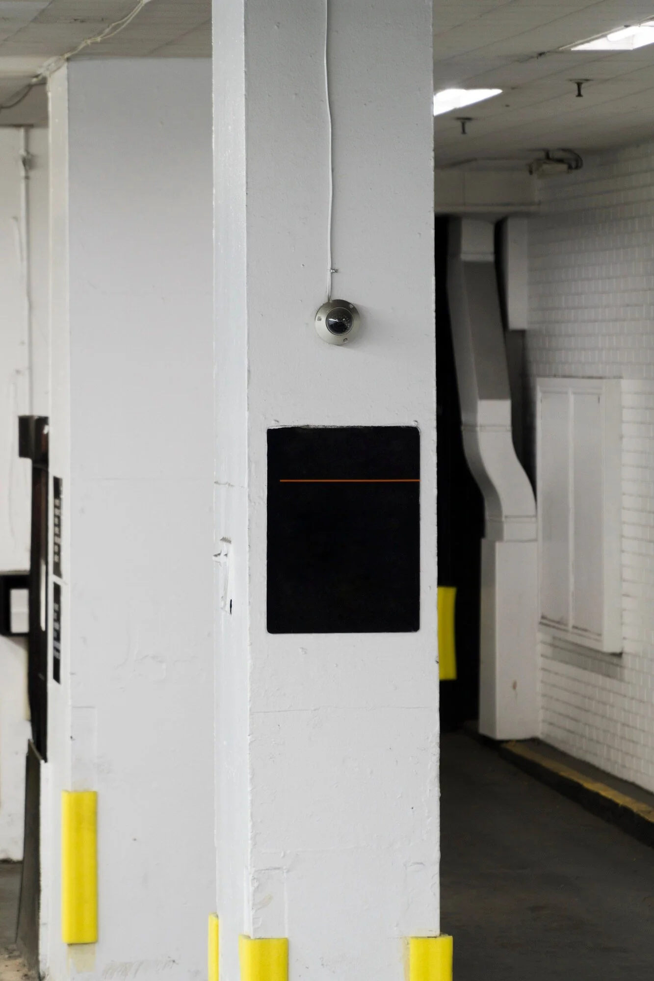 White concrete pillar with a security camera and a black square panel, in an indoor parking garage with yellow safety padding on the bottom of the pillar.