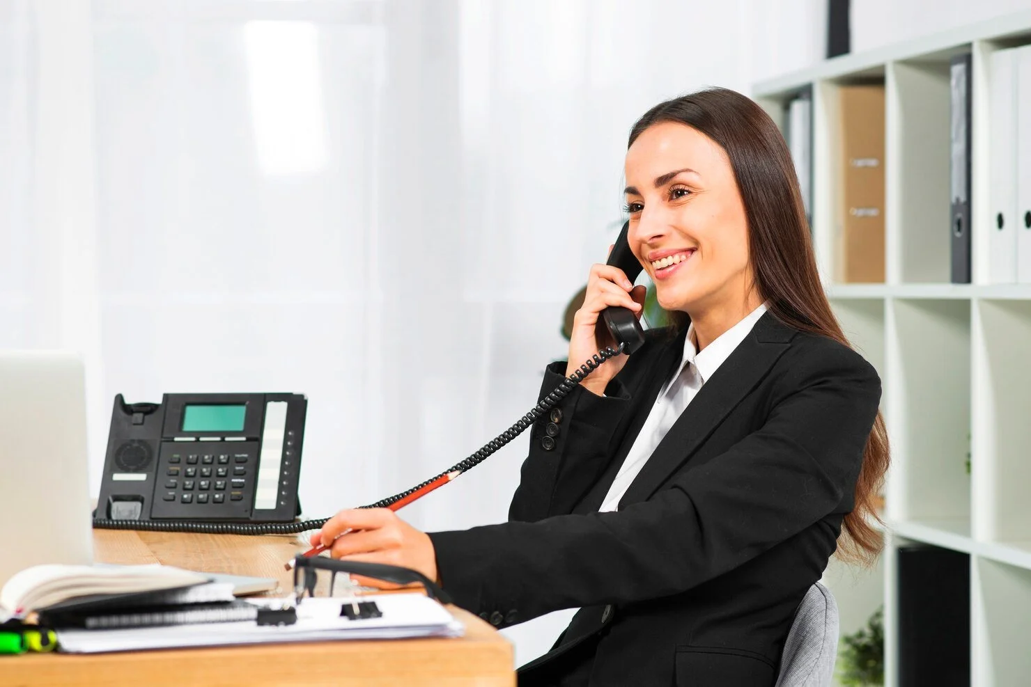 A woman in a black blazer with a white shirt, smiling on the phone in an office, with a desk and bookshelf behind her.