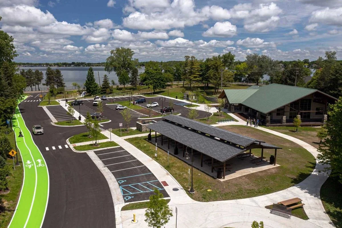 Aerial view of a park with a lake in the background, featuring a parking lot, a park shelter with a roof, a building with a green roof, a designated bike lane and pedestrian pathways, trees, and people walking.