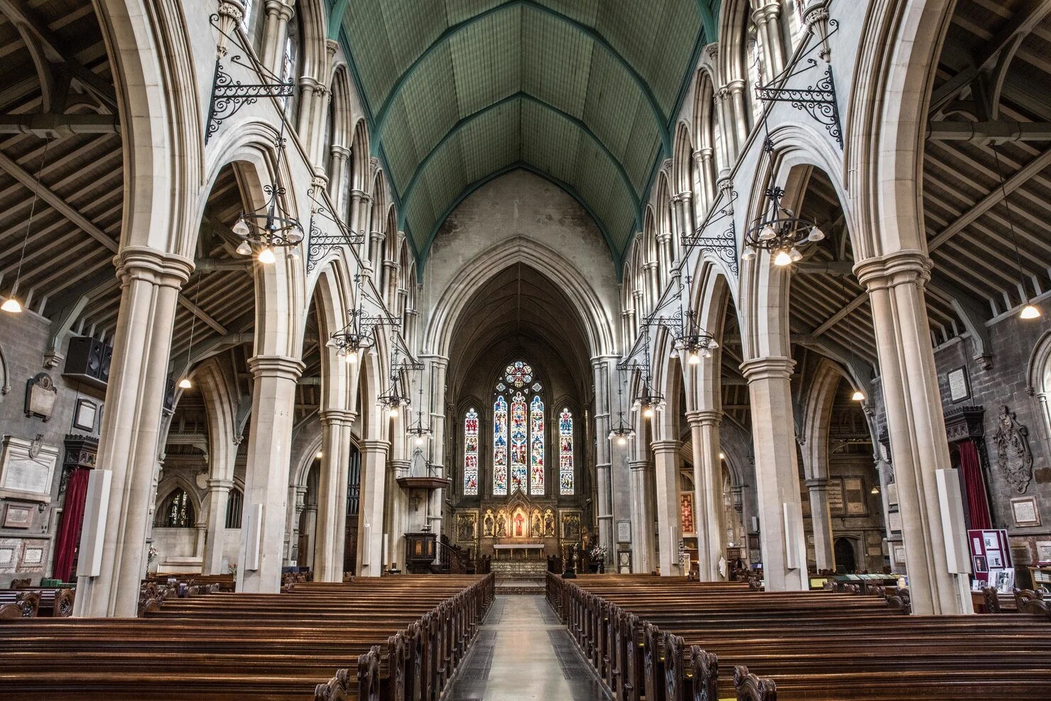 Interior of a historic church with high vaulted ceilings, stone columns, wooden pews, and stained glass windows at the altar.