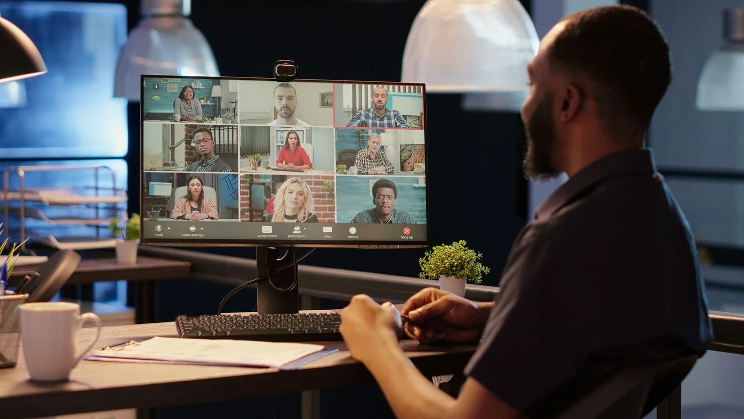 Man with beard participating in a virtual video conference at his desk with multiple meeting attendees on computer screen.