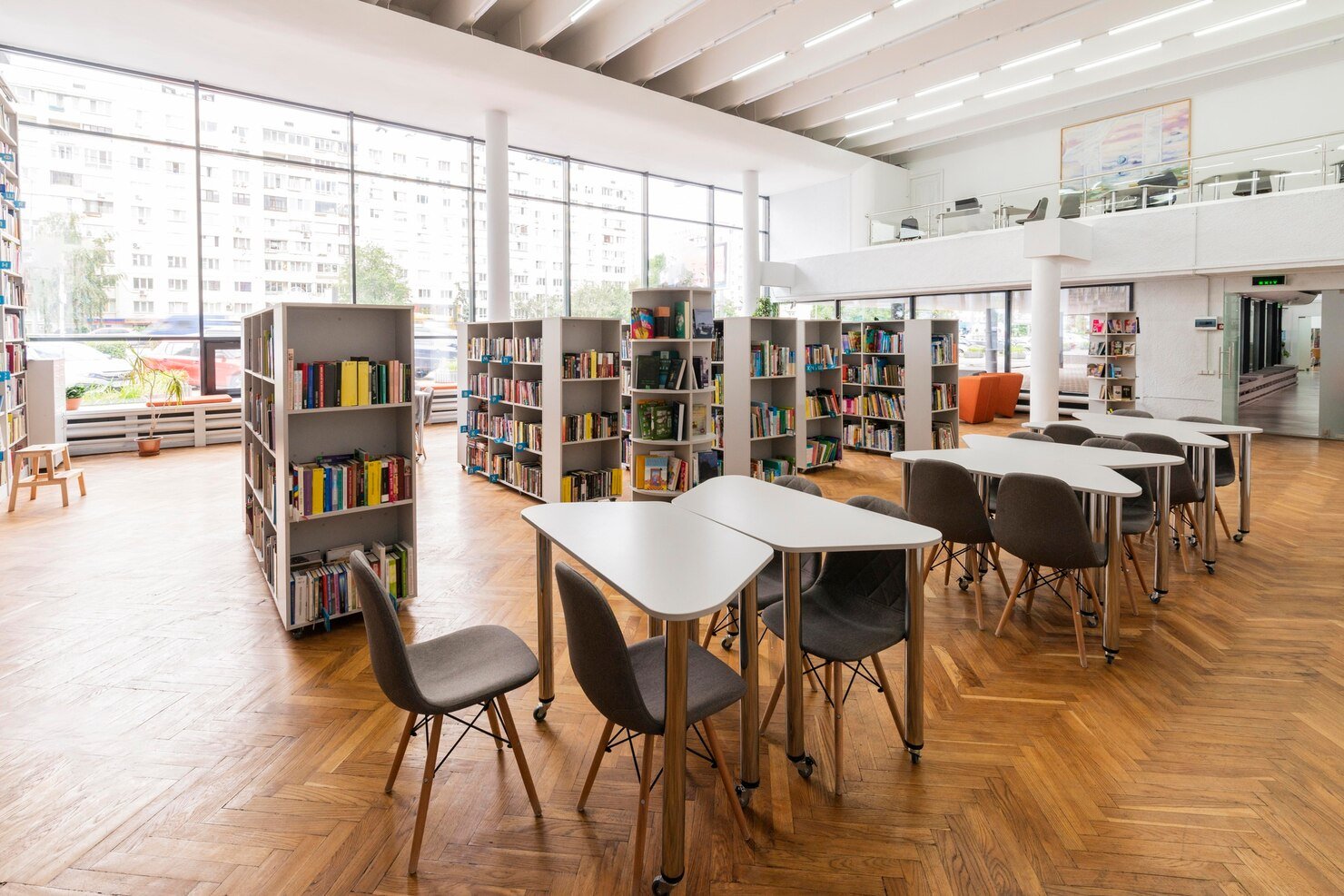 A modern, well-lit library interior with multiple bookshelves filled with books, several white tables, and gray chairs on a wooden floor. Large windows allow natural light to fill the space.