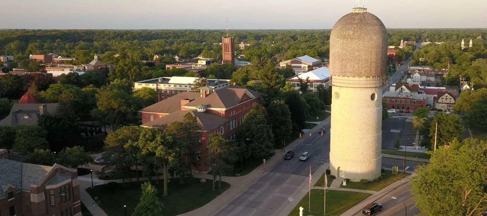 An aerial view of a small town with a prominent white cylindrical historic water tower on a corner, surrounded by trees, roads, cars, and brick buildings, with a mix of dense greenery and urban structures under a partly cloudy sky.