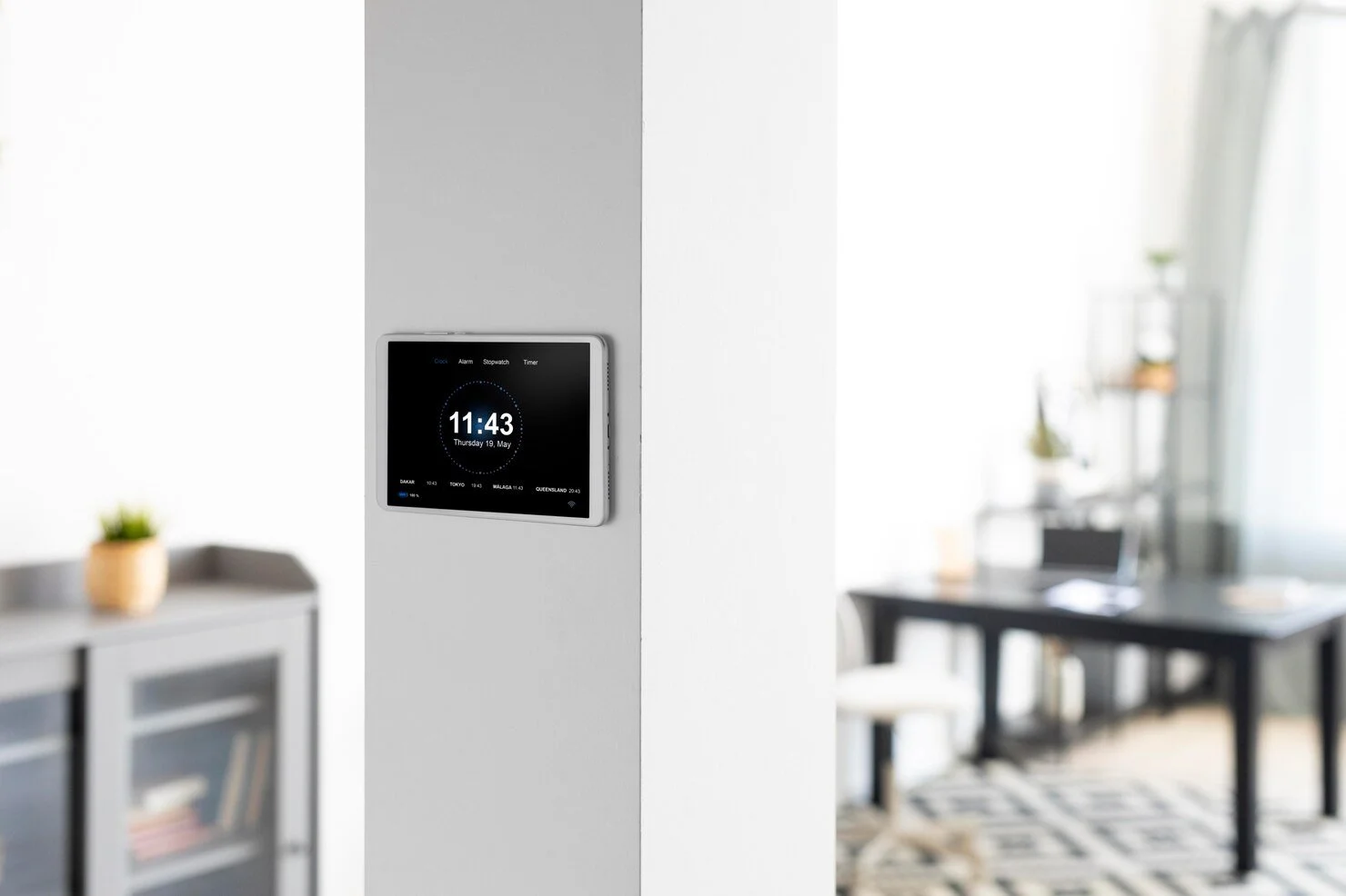 A digital clock on a white wall shows the time 11:43 on Thursday, May 19. The background includes a blurred modern, well-lit dining room with a black table, white chairs, and shelves with plants and decor.