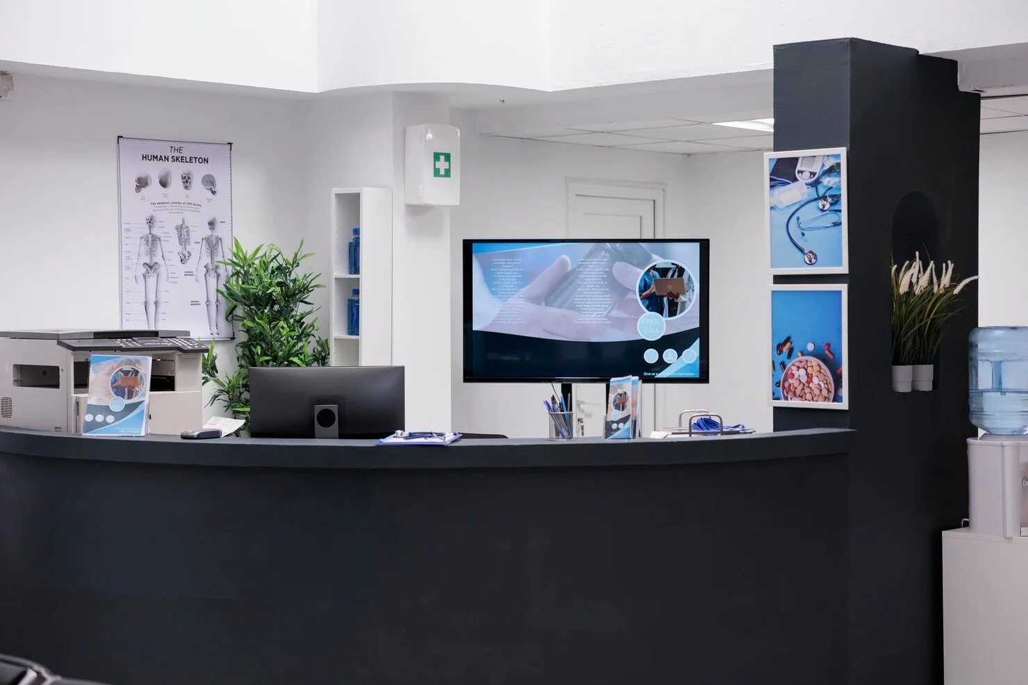 Reception desk in a medical office with a large screen monitor, medical posters, water cooler, and medical supplies.