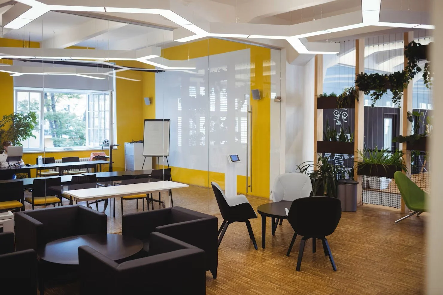 Modern open-concept office space with yellow accent walls, large windows, black and white furniture, and various potted plants.