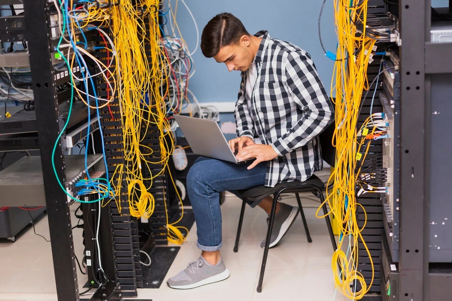 A man sitting in a server room working on a laptop amidst tangled yellow and blue network cables.