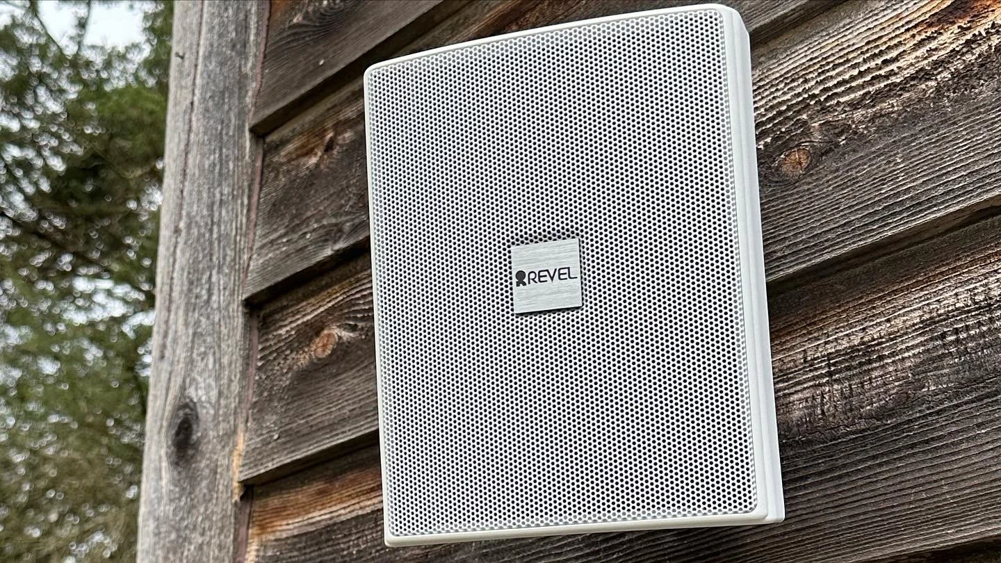 A white REVEL outdoor speaker mounted on weathered wooden siding of a building with trees visible in the background.