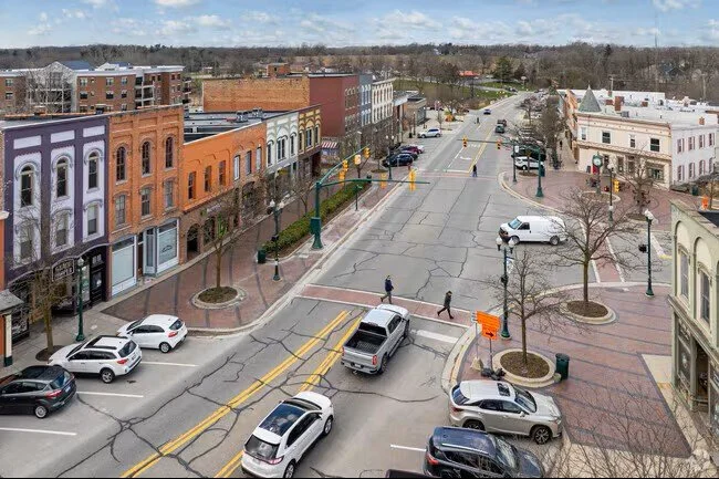 Aerial view of a small-town intersection with brick buildings, parked cars, pedestrians, and leafless trees in early spring or late winter.