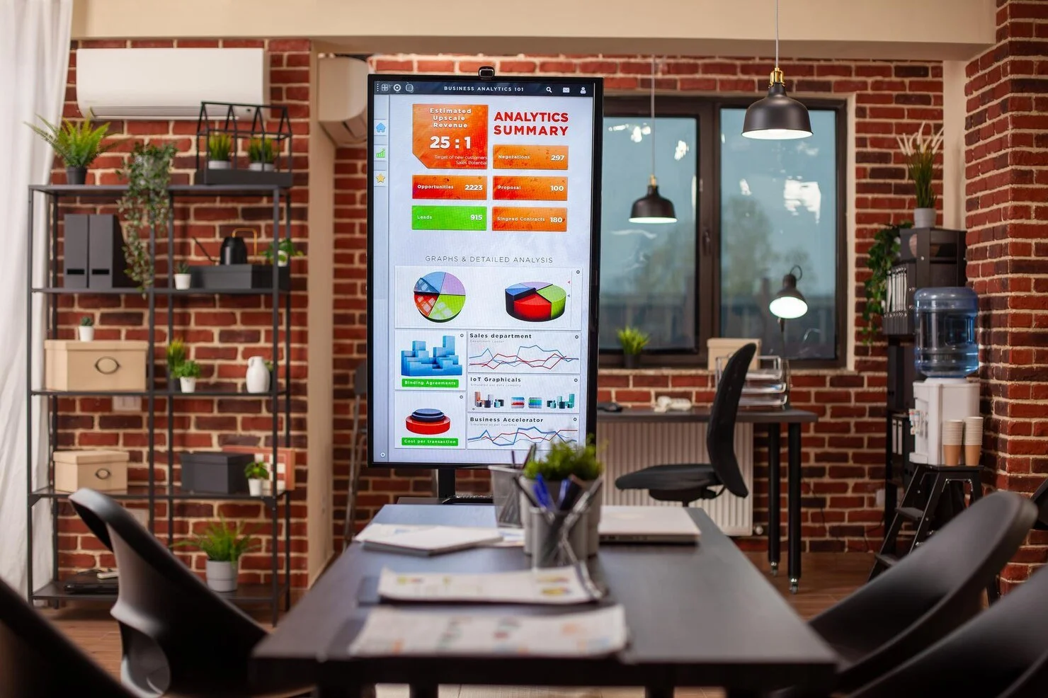 Office conference room with brick walls, a large monitor displaying data analytics, a table with papers and pens, chairs, shelves with plants, a water dispenser, and windows with view outside.
