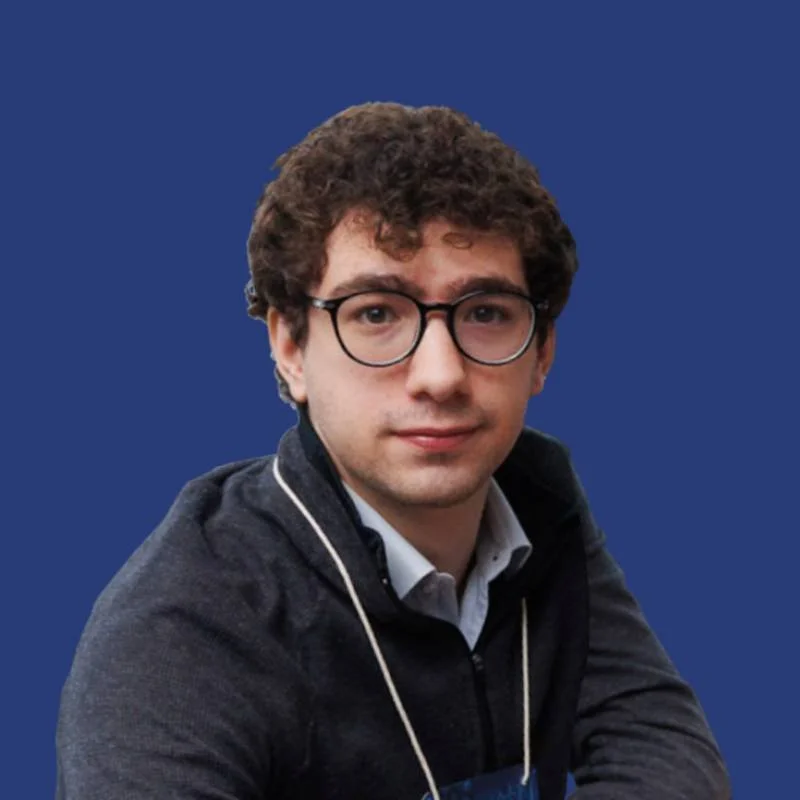 Jovem adulto de cabelo cacheado, usando óculos de armação preta, camiseta azul clara com colarinho e expressão neutra, em ambiente com teto de LEDs e luminária arredondada no fundo.