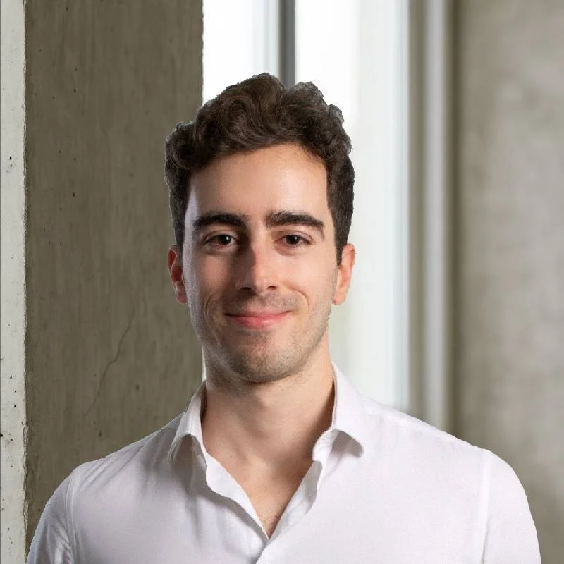 Homem sorridente usando camisa branca, posando ao lado de uma parede de concreto, com janela ao fundo.