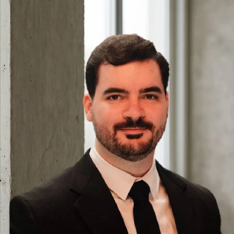Homem de terno preto, camisa branca e gravata preta, com barba e cabelo escuro, posando em ambiente interno com parede de concreto ao lado.