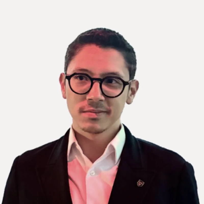 Homem com cabelo castanho, óculos de armação preta, camisa branca e blazer preto, olhando para o lado, na uma sala com parede clara e janela ao fundo.